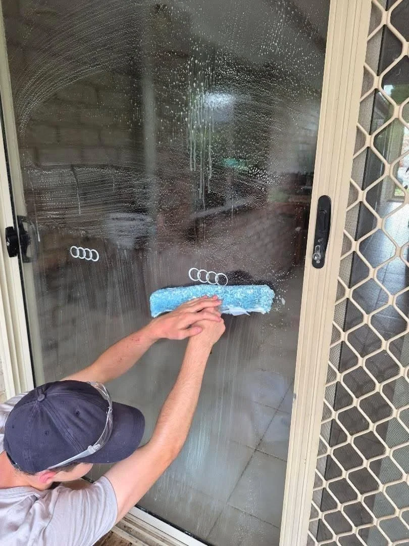 Person cleaning a business's glass sliding door with a blue sponge or cloth, soap and water creating suds on the glass. The cleaning man cleans home's windows.