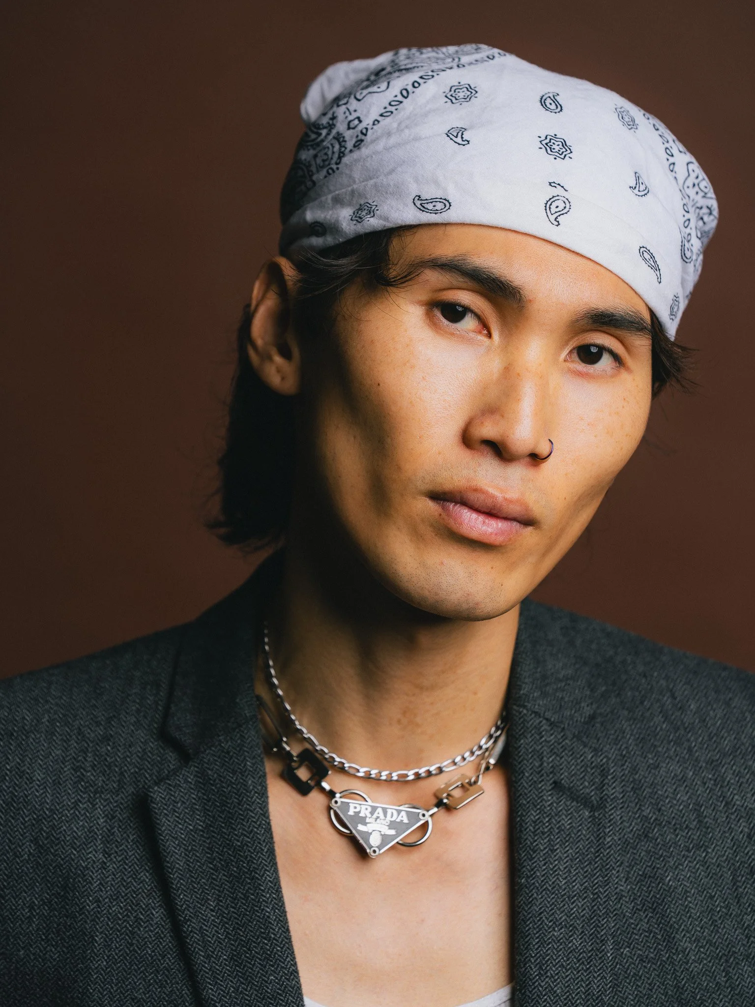 A young person with light skin and dark hair wearing a white bandana, layered silver necklaces including one with a Prada logo, a septum ring, and a dark blazer, posing against a brown background.