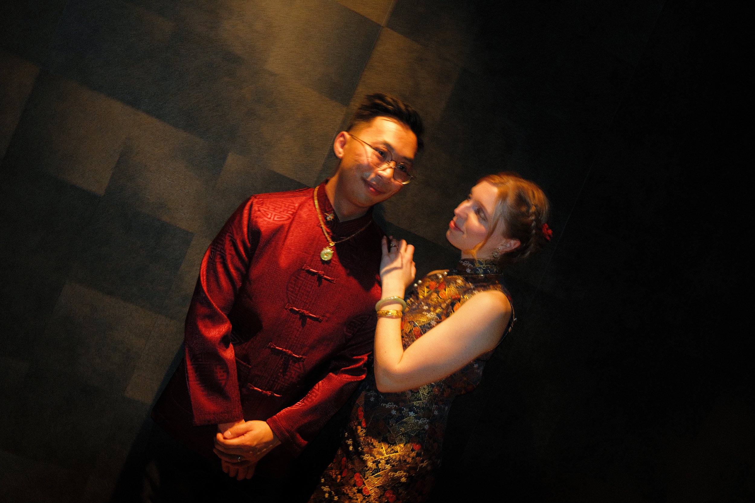 A man and woman holding hands and smiling at each other, dressed in traditional Asian attire, standing close in a dimly lit space.