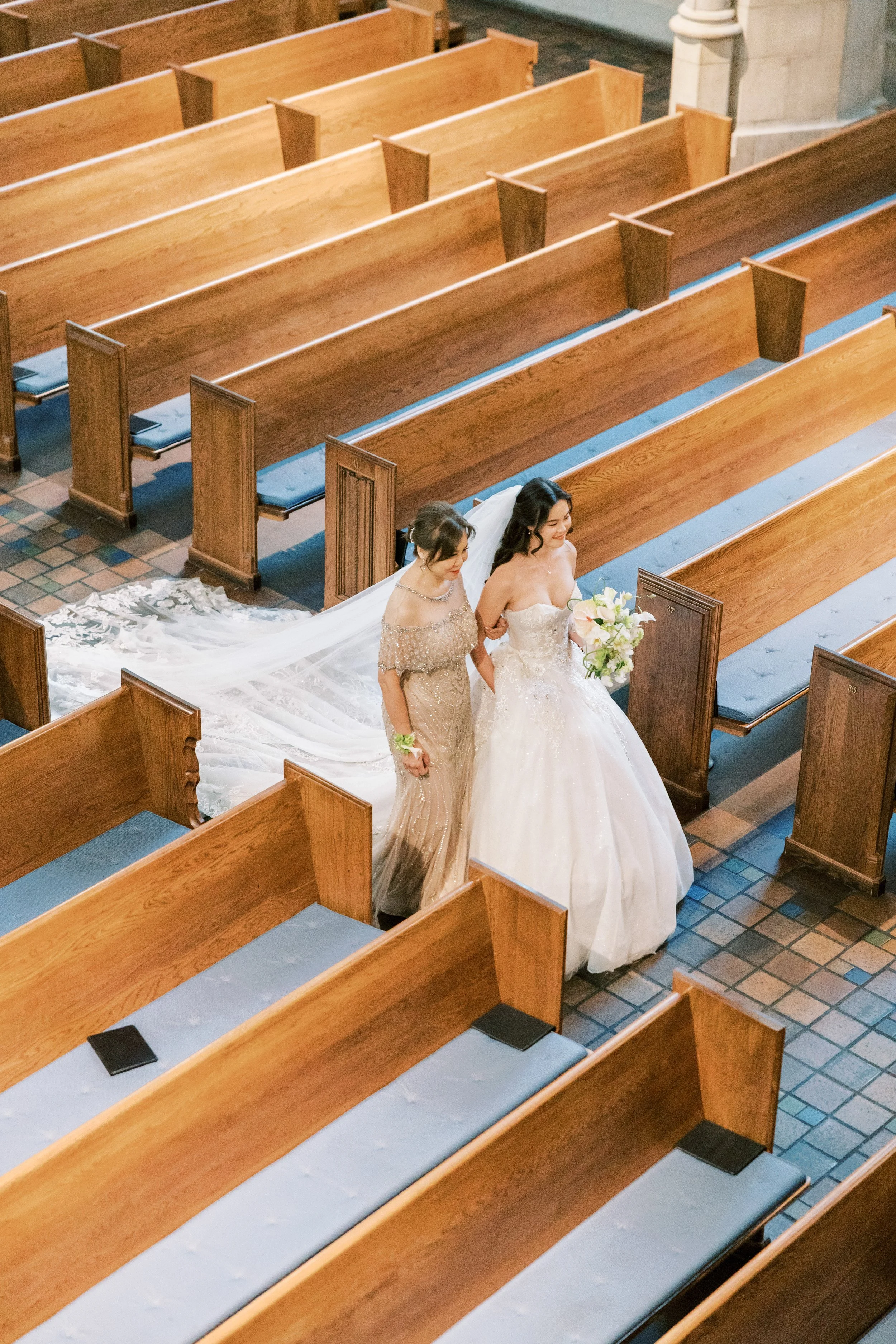 Two women, dressed in wedding attire, walking down the aisle of a church with wooden pews and tiled floor. One is a bride holding a bouquet, accompanied by another woman in a gown.