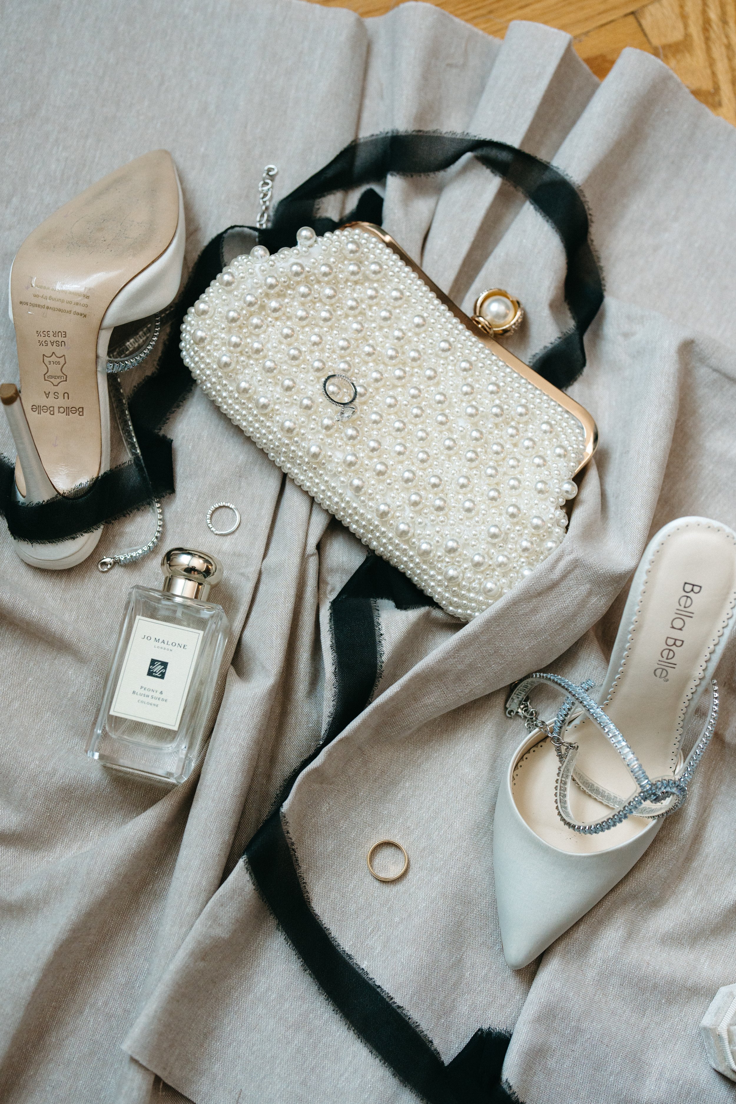 A collection of jewelry, a small pearl-embellished clutch purse, a pair of white shoes with rhinestone ankle straps, a perfume bottle, and a few rings arranged on a fabric surface.