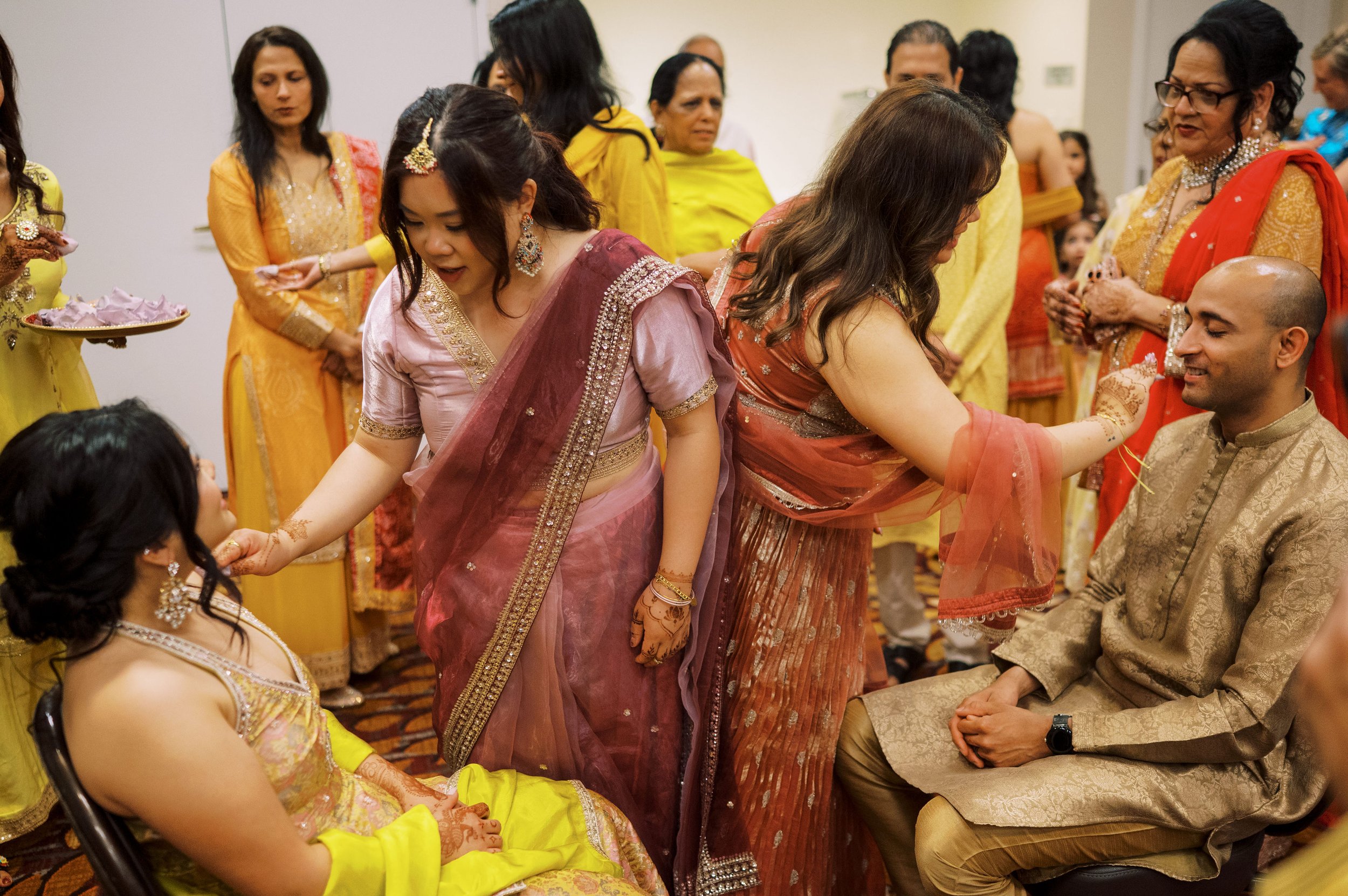 Women dressed in colorful traditional Indian attire participating in a ceremony, with some kneeling and others standing, in an indoor setting.