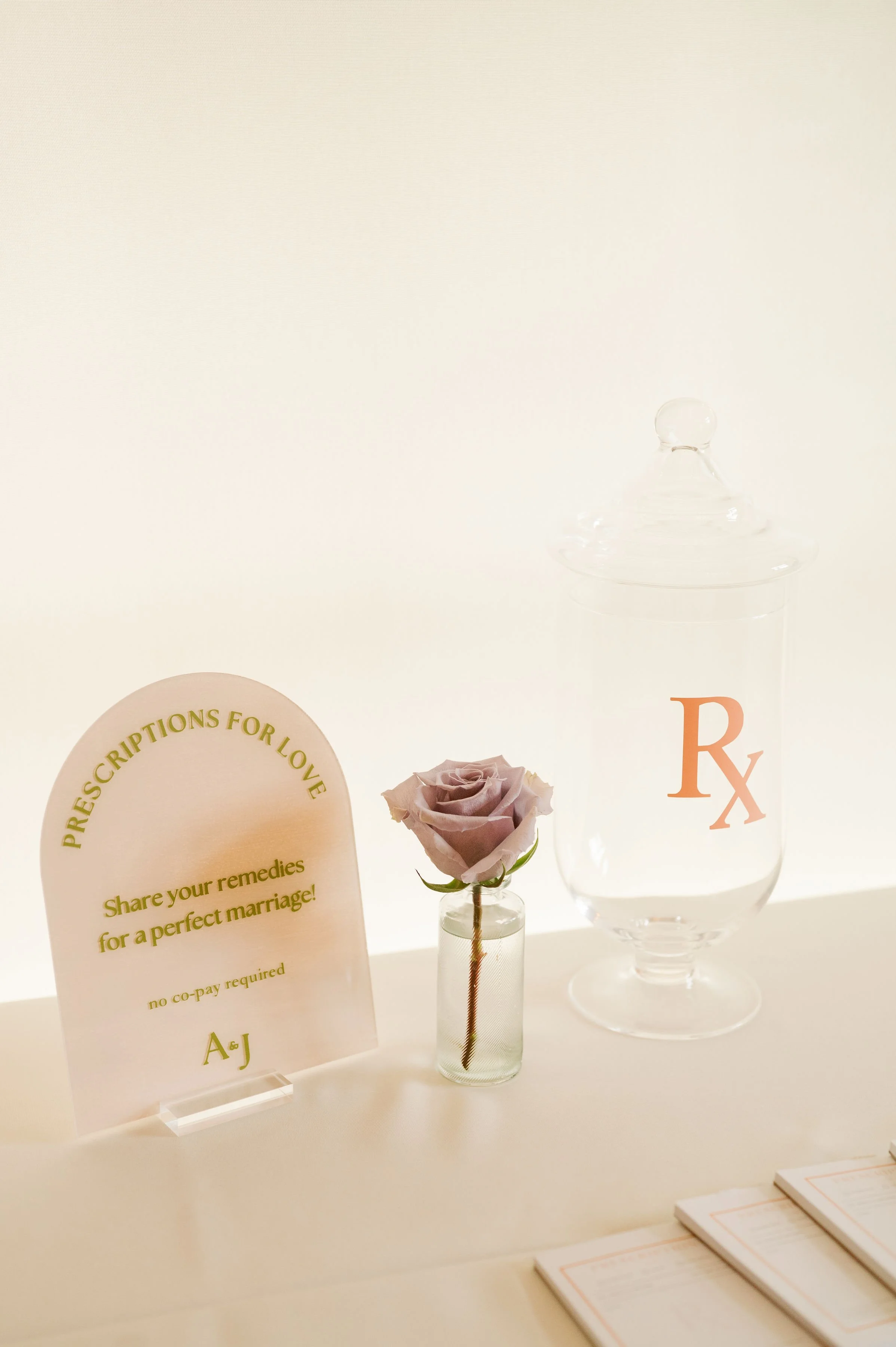 A small glass vase with a single pink rose, a sign about prescriptions for love, a glass of water with an orange Rx symbol label, and some pamphlets on a white surface with a light background.