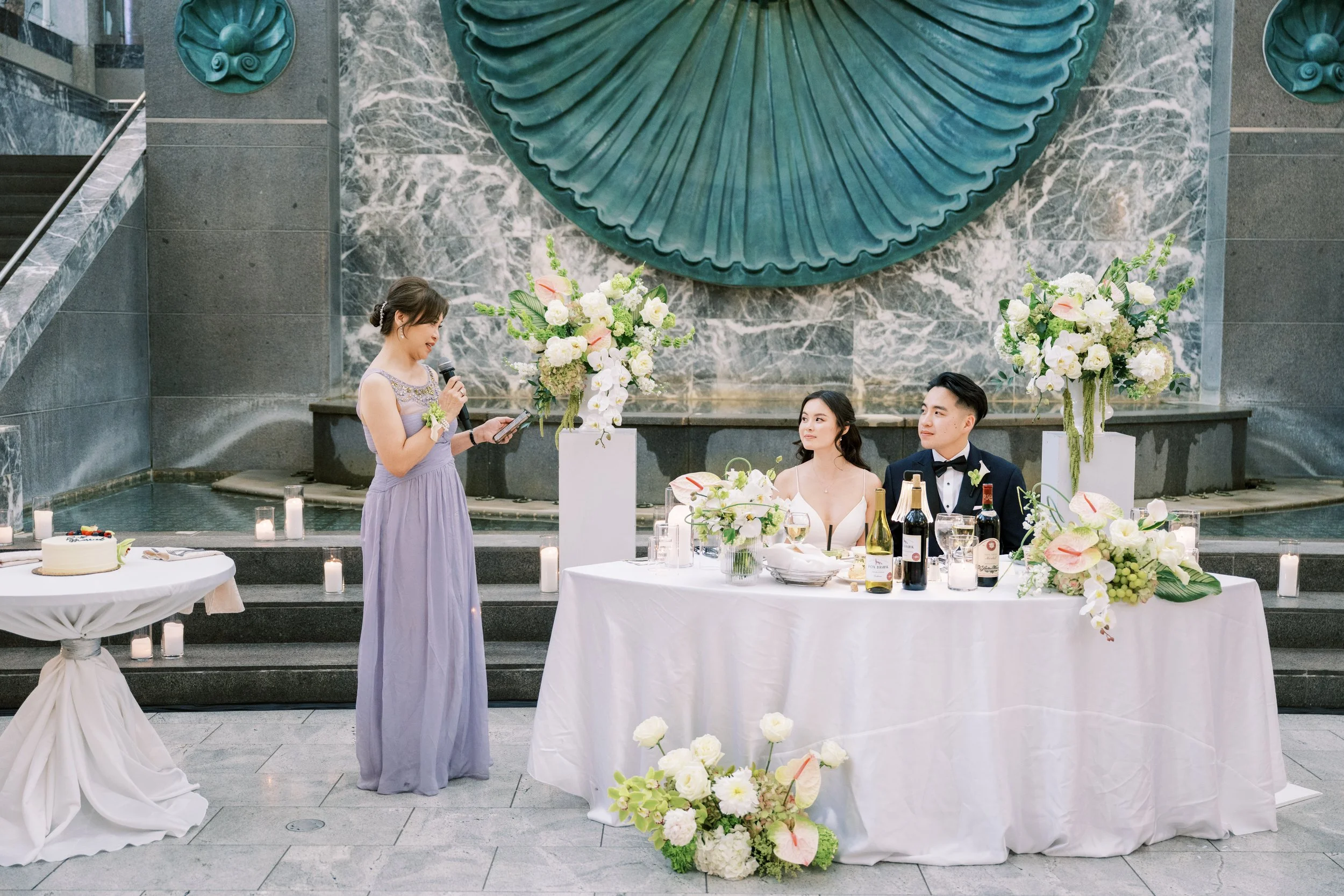 A woman giving a speech at a wedding reception with a bride and groom sitting at a decorated table. The setting features large floral arrangements and a blue decorative wall piece.