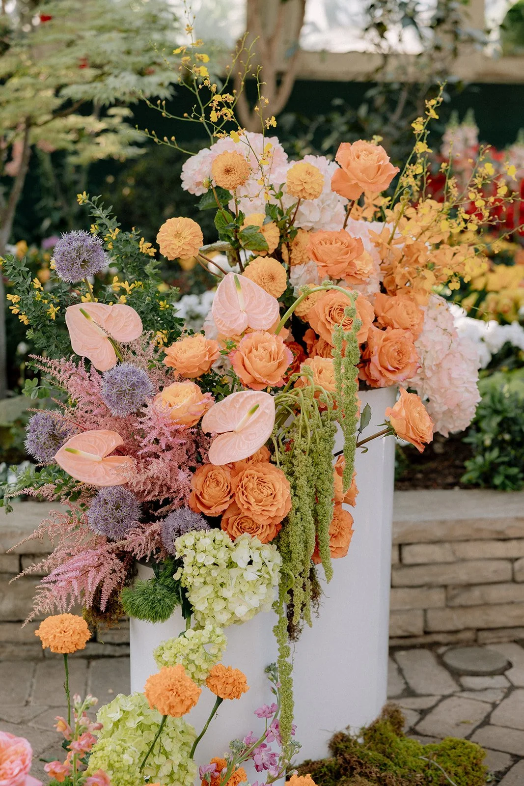 A vibrant floral arrangement in a white vase, featuring peach roses, purple alliums, pink anthuriums, and various greenery, set outdoors on a stone surface.