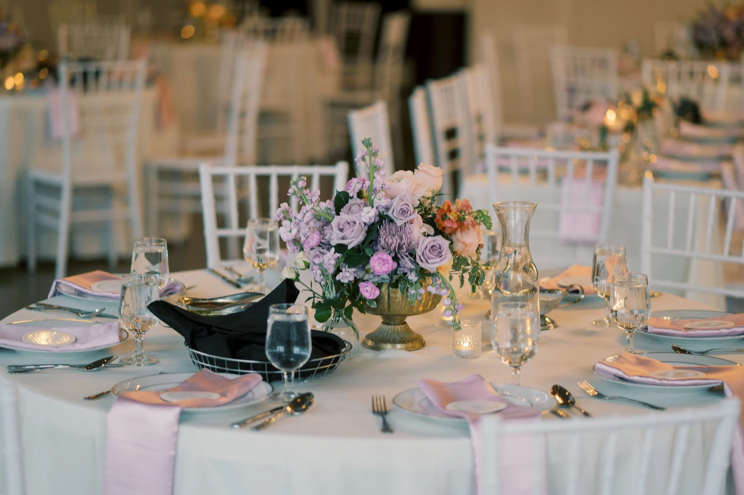 Elegant banquet table decorated with a floral centerpiece featuring pink, purple, and cream-colored flowers, surrounded by glasses, plates, and silverware, set for a formal event in an indoor venue.