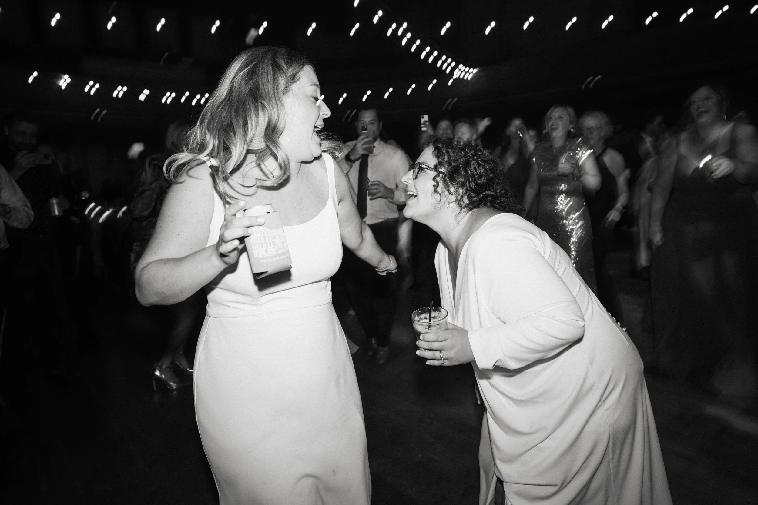 Two women in white dresses sharing a joyful moment at a formal event, with other people dancing and taking photos in the background.