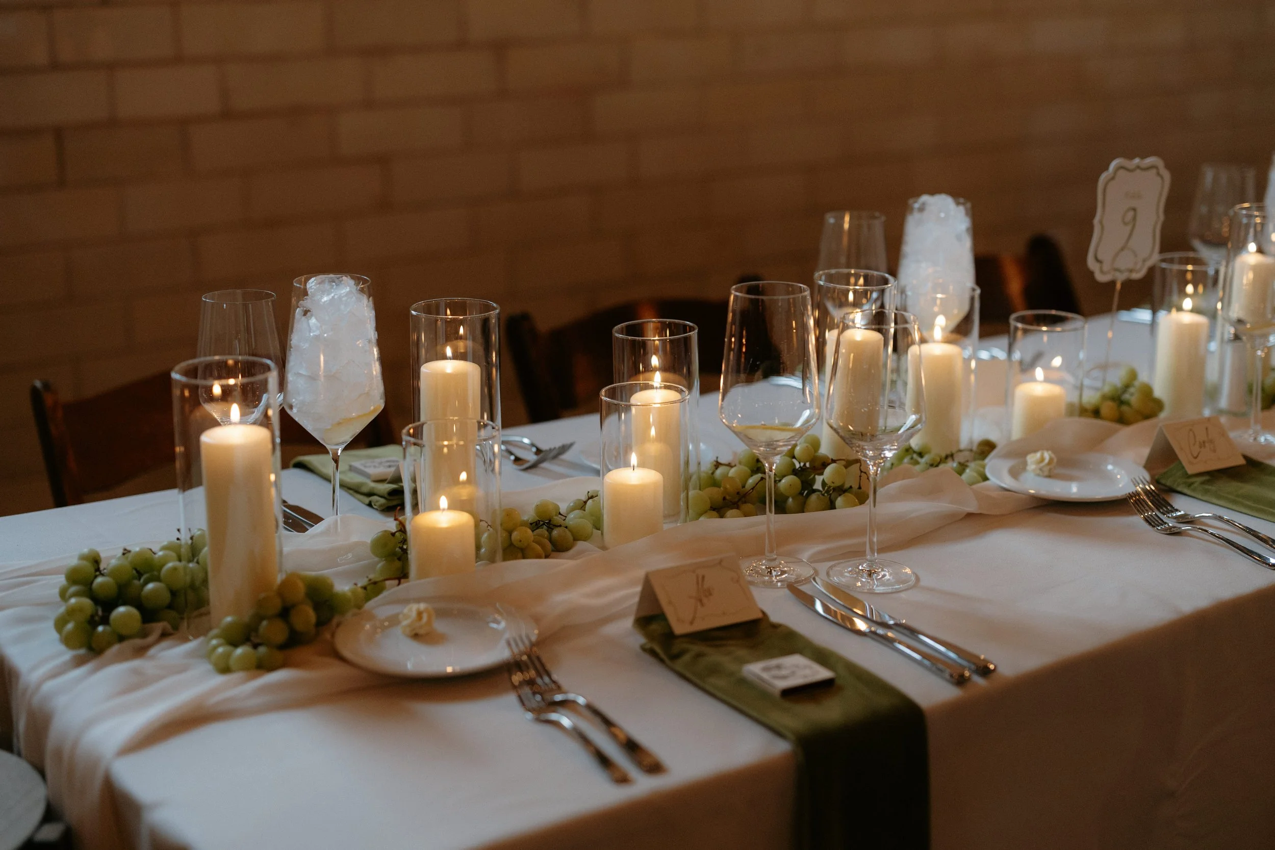 A decorated dining table with white candles, white grapes, wine glasses, and place settings in a warmly lit room with a brick wall background.