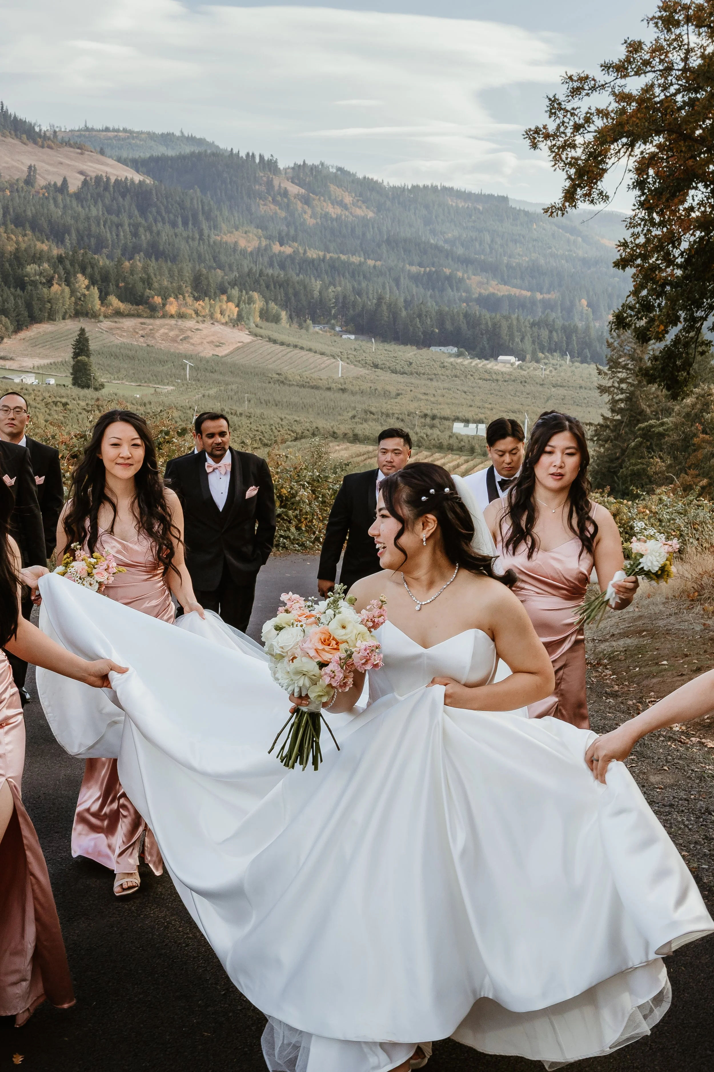 A bride in a white wedding gown holding a bouquet of flowers and smiling, surrounded by bridesmaids in pink dresses and groomsmen in black tuxedos, outdoors with a scenic mountain landscape in the background.