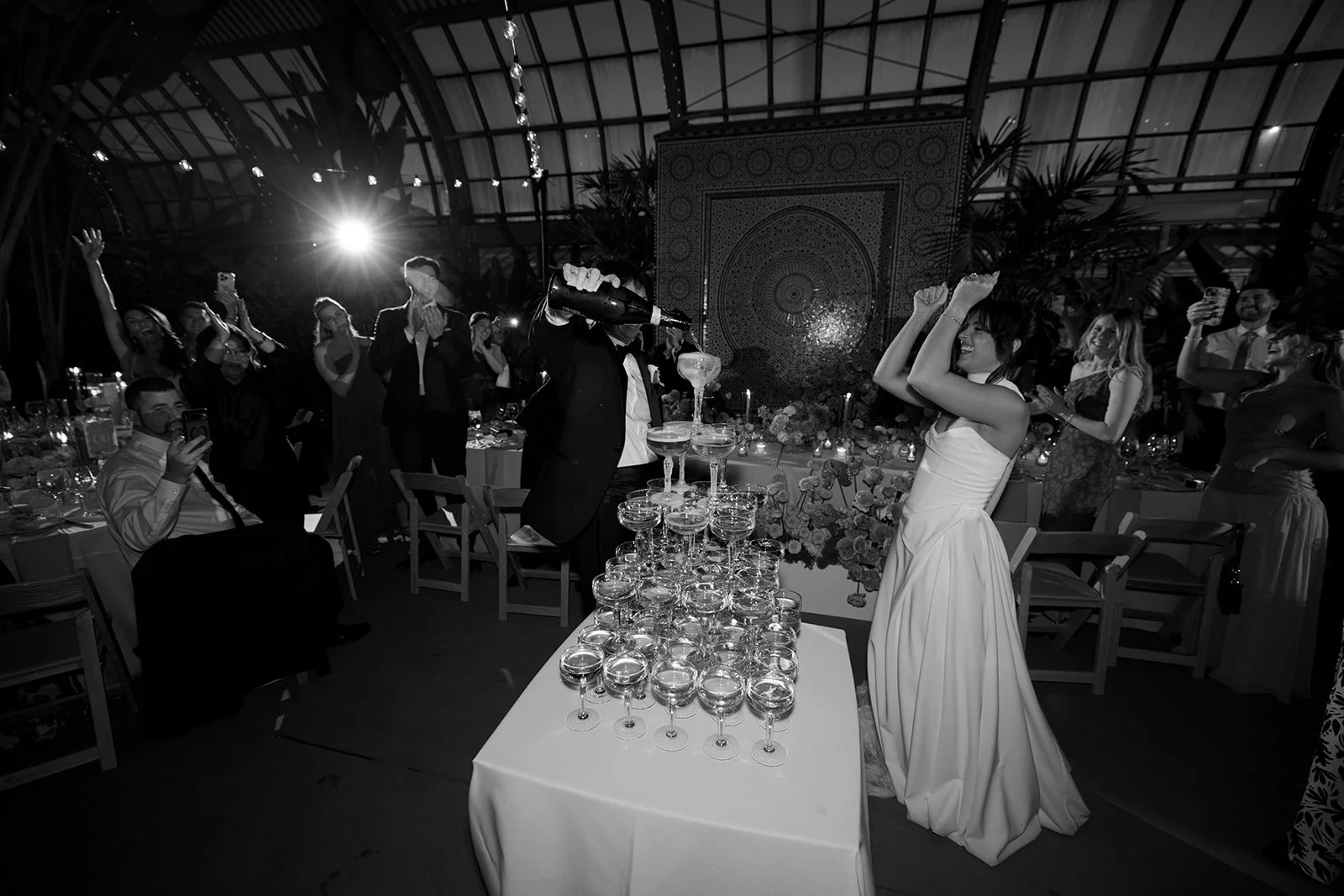 Black and white photo of a wedding celebration with a bride in a strapless white gown dancing, a groom pouring champagne into a tower of glasses, and guests applauding and taking photos in a large indoor venue with a decorated backdrop and floral arr