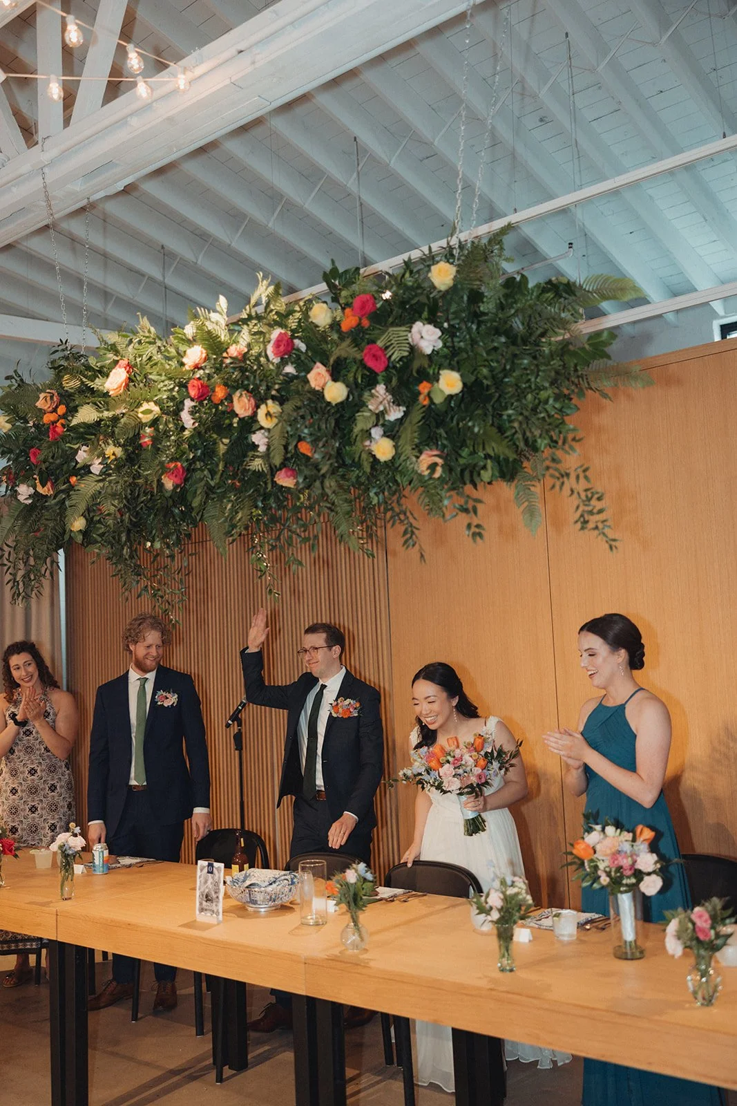 A wedding reception with a large floral arrangement hanging above a head table where five people are standing, including a bride in a white dress and a groom in a dark suit. They are all smiling and clapping or raising their hands, with wooden panels