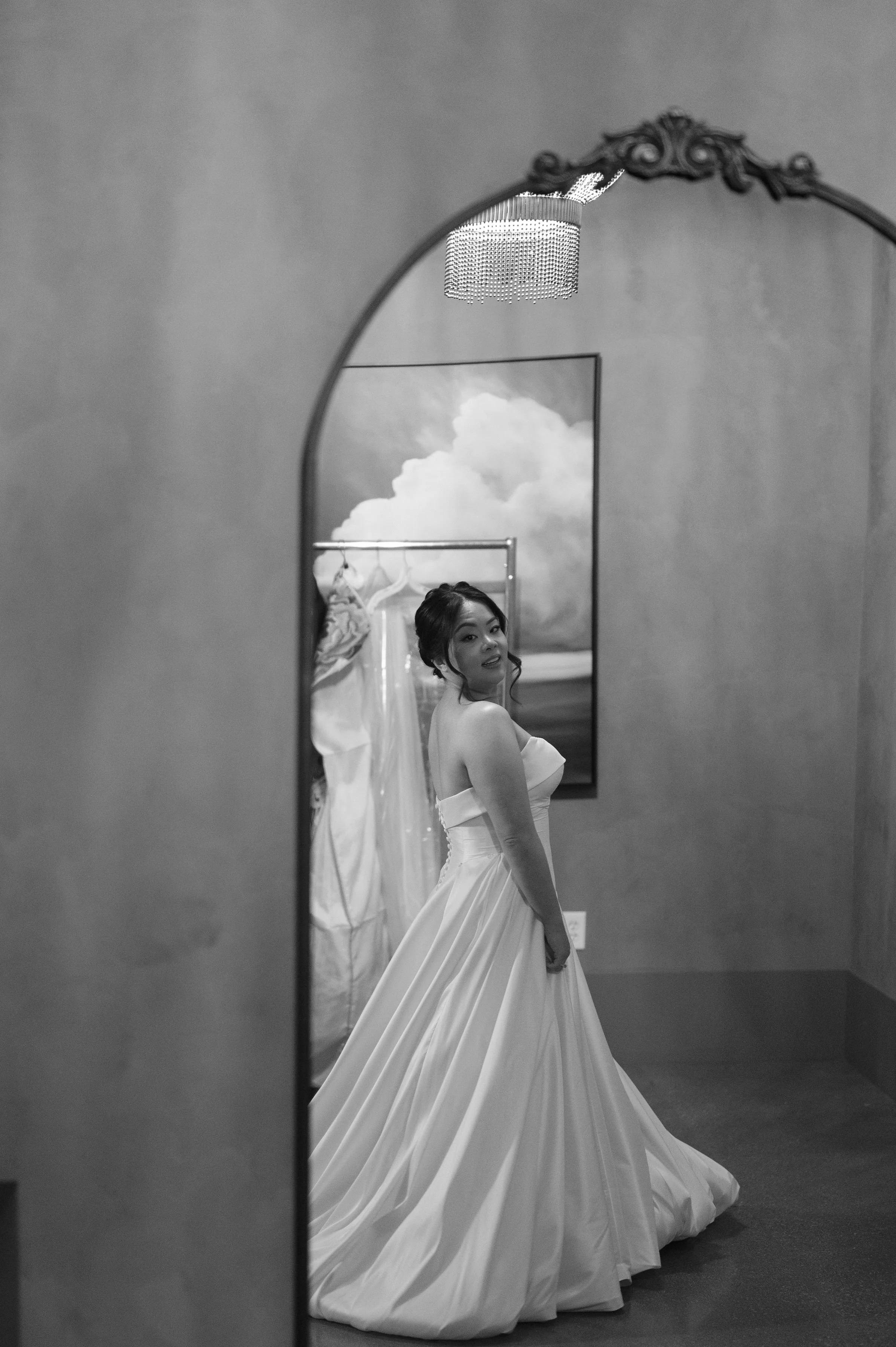 A woman in a wedding dress reflected in a mirror, with a cloudy sky painting behind her, and a chandelier hanging from the ceiling.