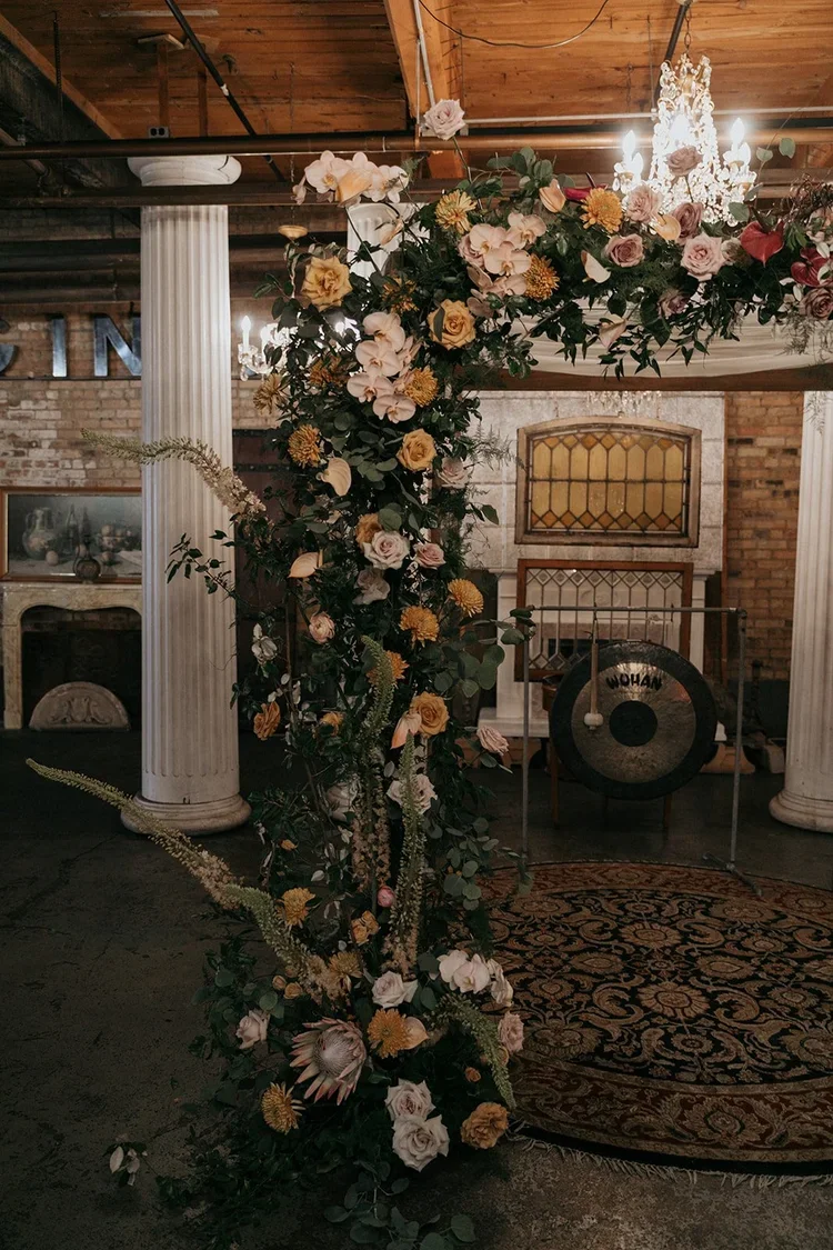 A floral wedding arch with white, pink, and yellow flowers and greenery, set in an indoor venue with brick walls, white pillars, a chandelier, and a rug, with a gong in the background.