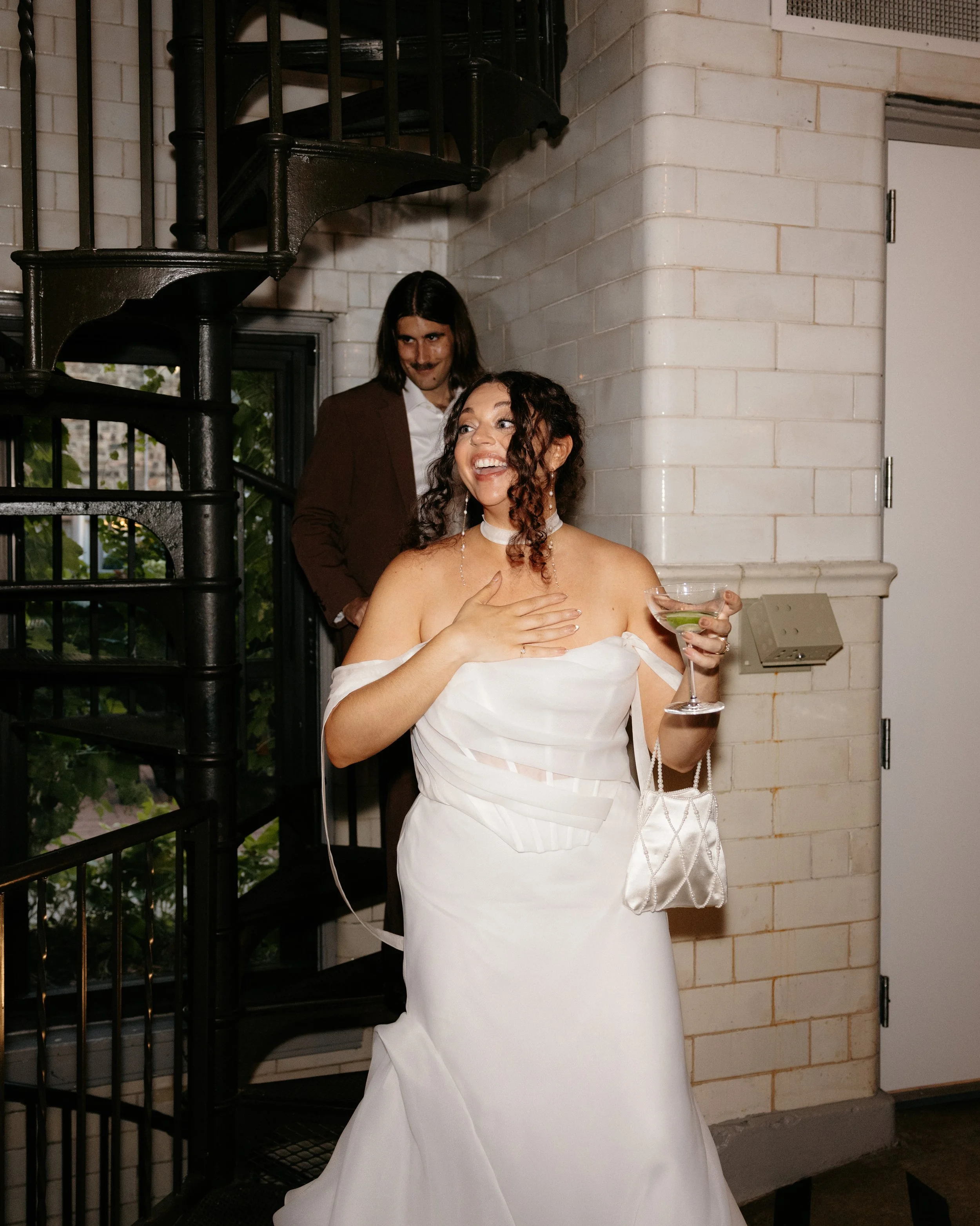 A woman in a white dress holding a martini glass, smiling and looking surprised, with a man in a brown suit with long hair and a beard in the background, indoors near a white brick wall and black spiral staircase.