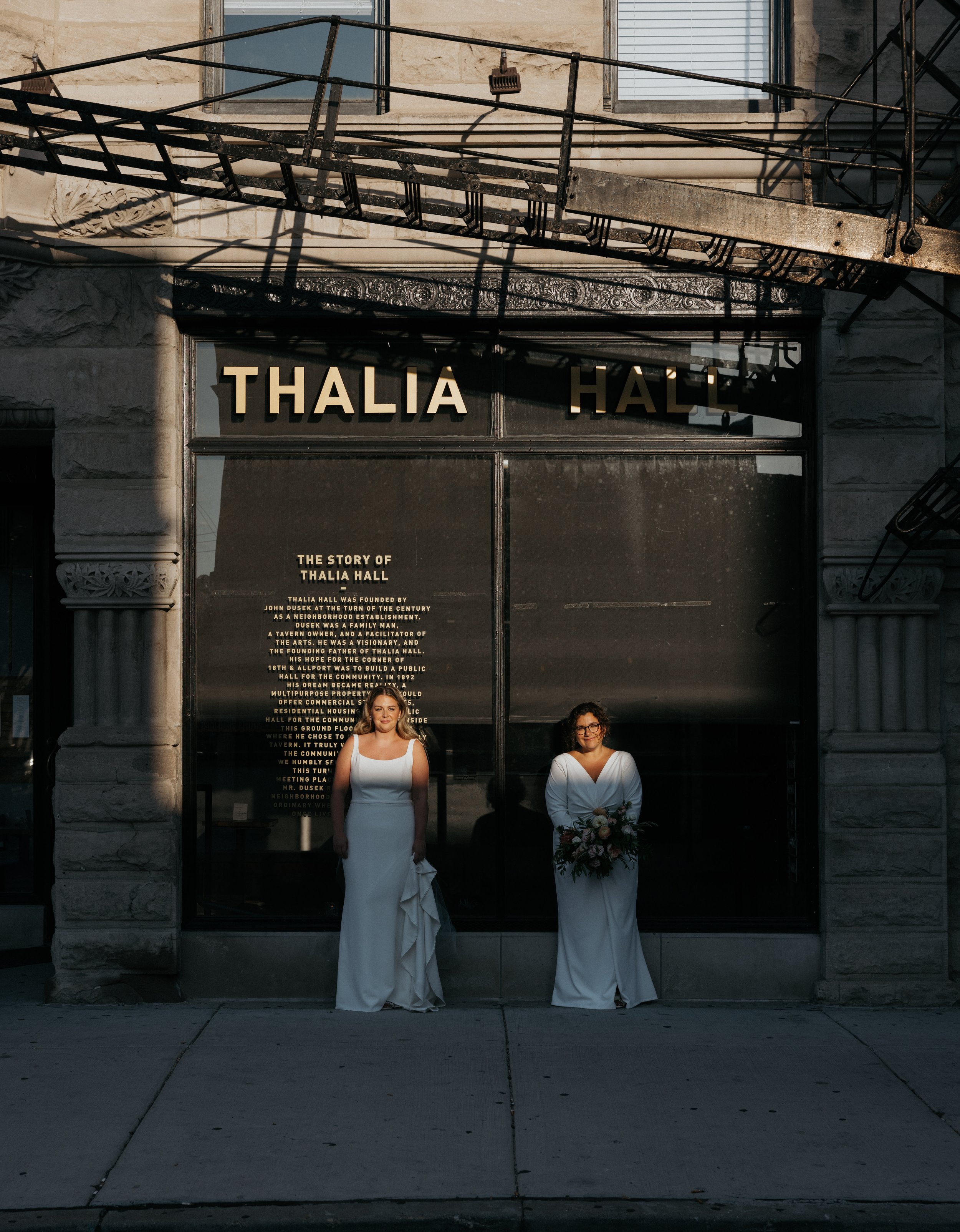 Two women in white wedding dresses stand outside of Thalia Hall with a brick facade. One woman holds a bouquet, and the other stands with her hands by her sides, in front of a glass entrance displaying the words "Thalia Hall" and a plaque describing 