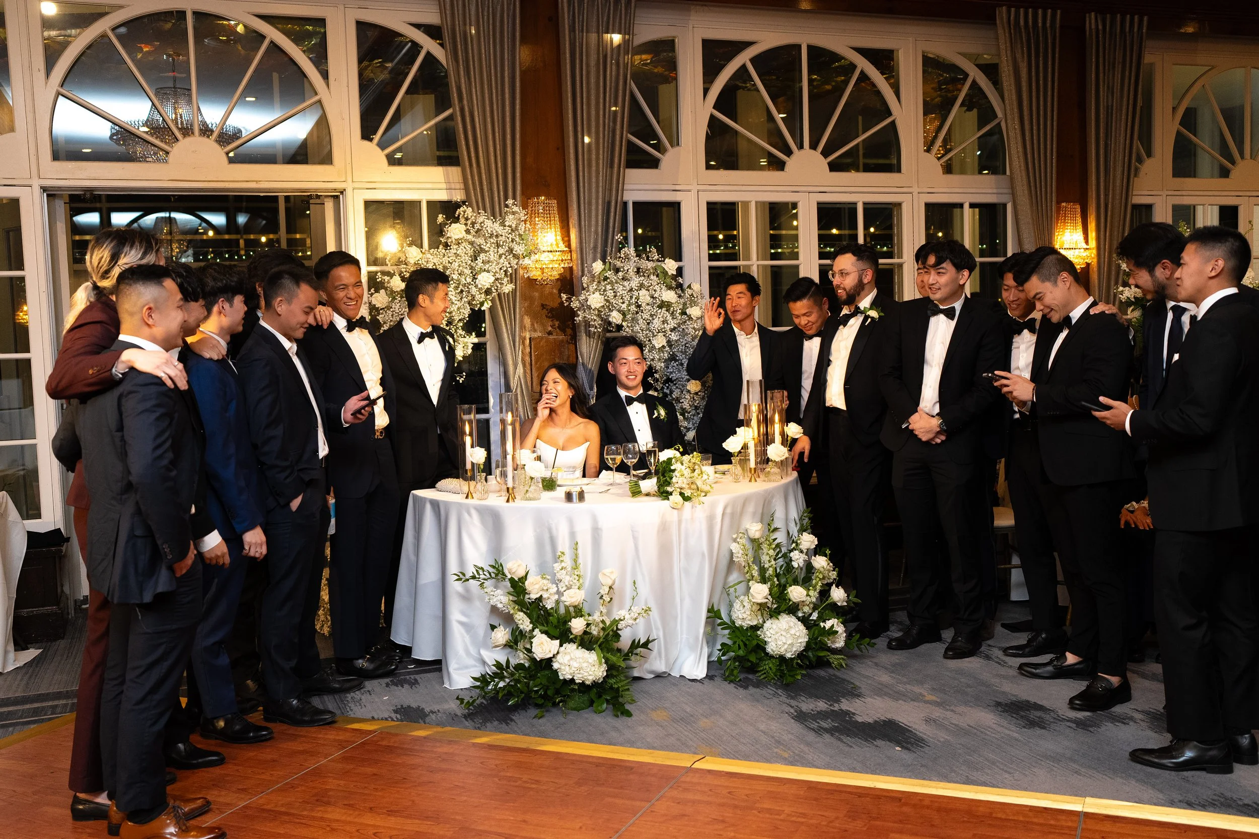 A wedding celebration with a group of men dressed in formal black tuxedos gathered around a bride and groom seated at a table. The scene is decorated with white flowers and elegant decor, inside a room with large arched windows and curtains, with war