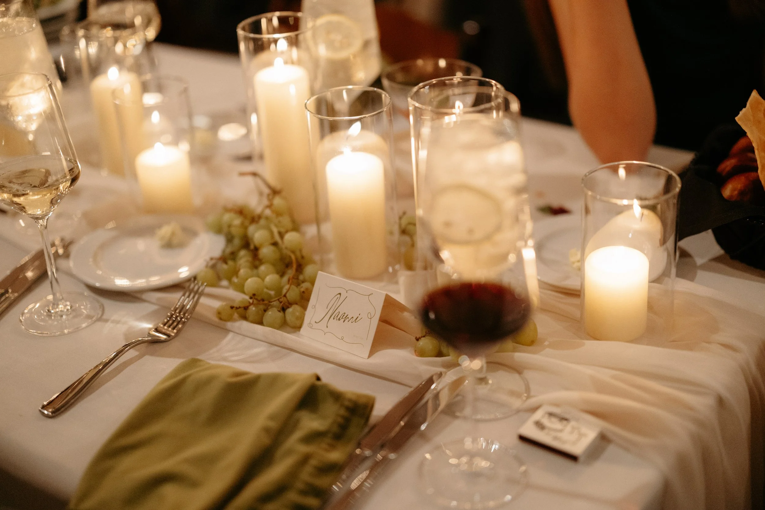 A formal dinner table decorated with lit candles, white grapes, wine glasses filled with white and red wine, silverware, and a place card with the name Naomi.