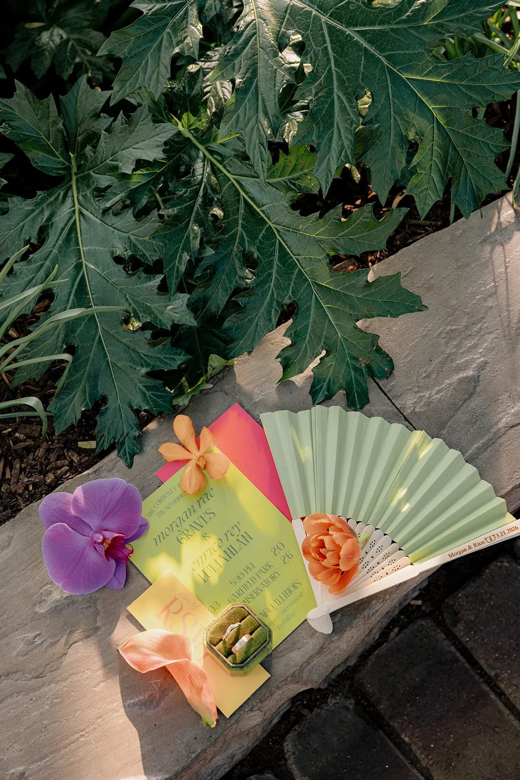 Colorful wedding invitation card with decorative flowers and a small box with rings, placed on a stone ledge beneath large green leaves.