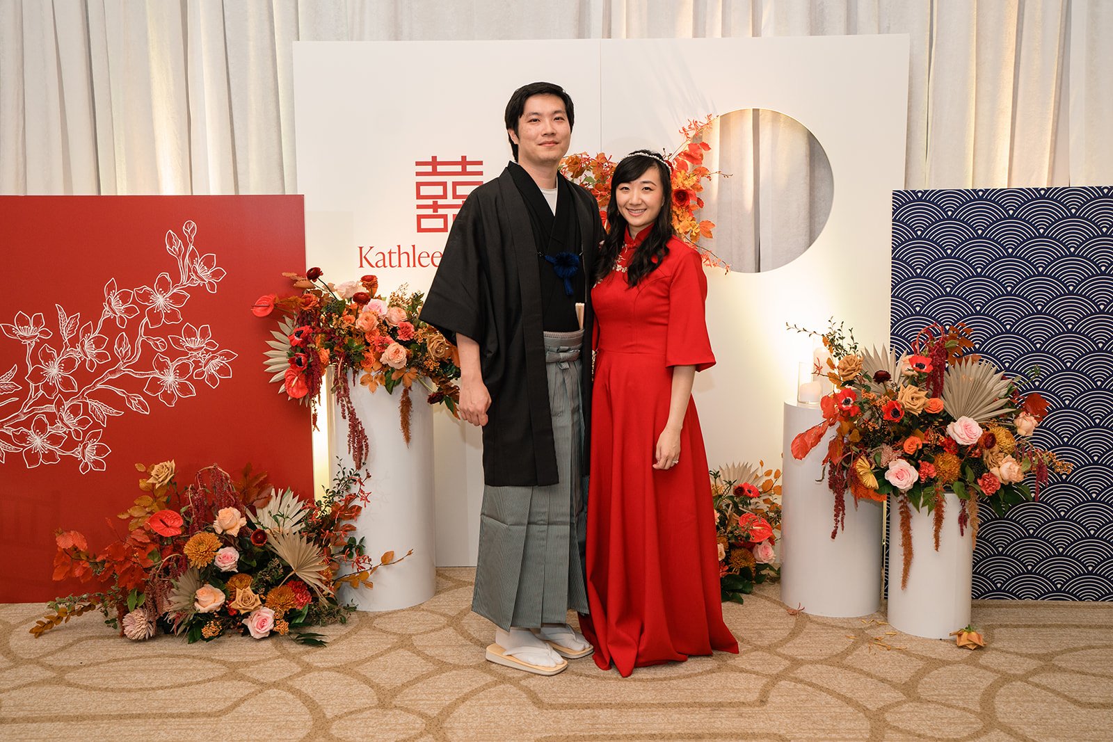 A couple dressed in traditional Japanese attire, standing among floral arrangements in a decorated setting. The man is wearing a black kimono with gray patterned hakama, and the woman is in a red kimono dress, smiling at the camera.