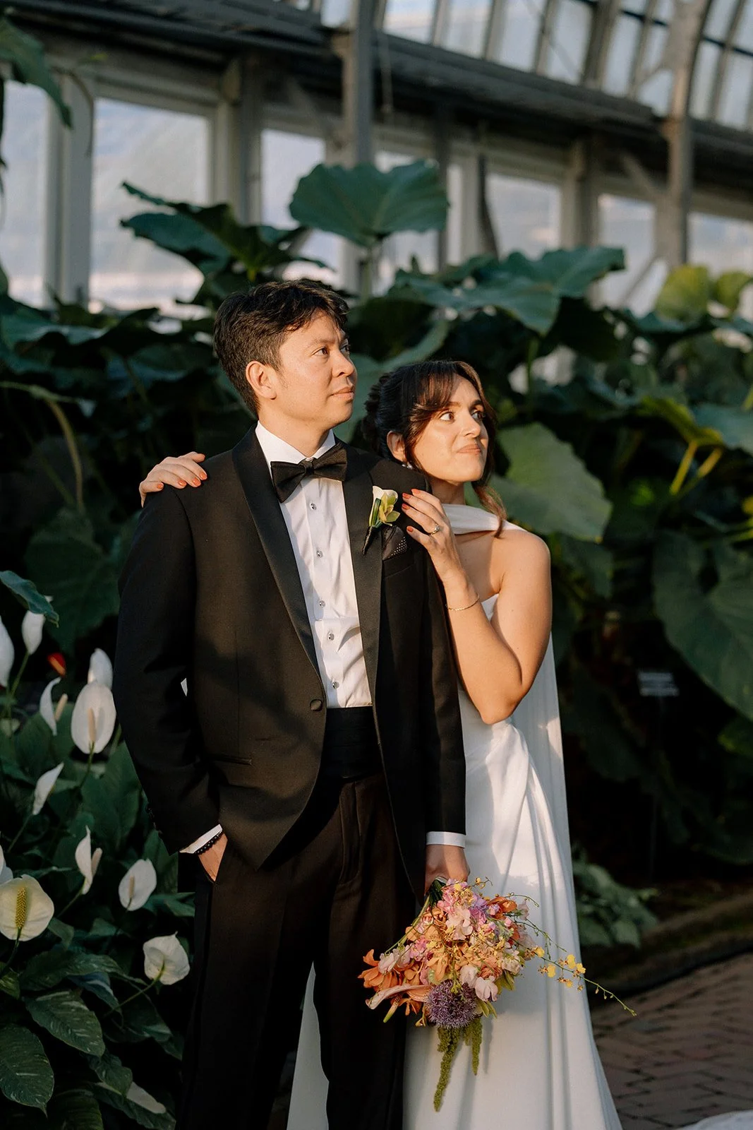 A couple dressed in wedding attire standing in a greenhouse with large green leaves, the man in a black tuxedo and the woman in a white dress holding a pink bouquet, both looking contemplatively.