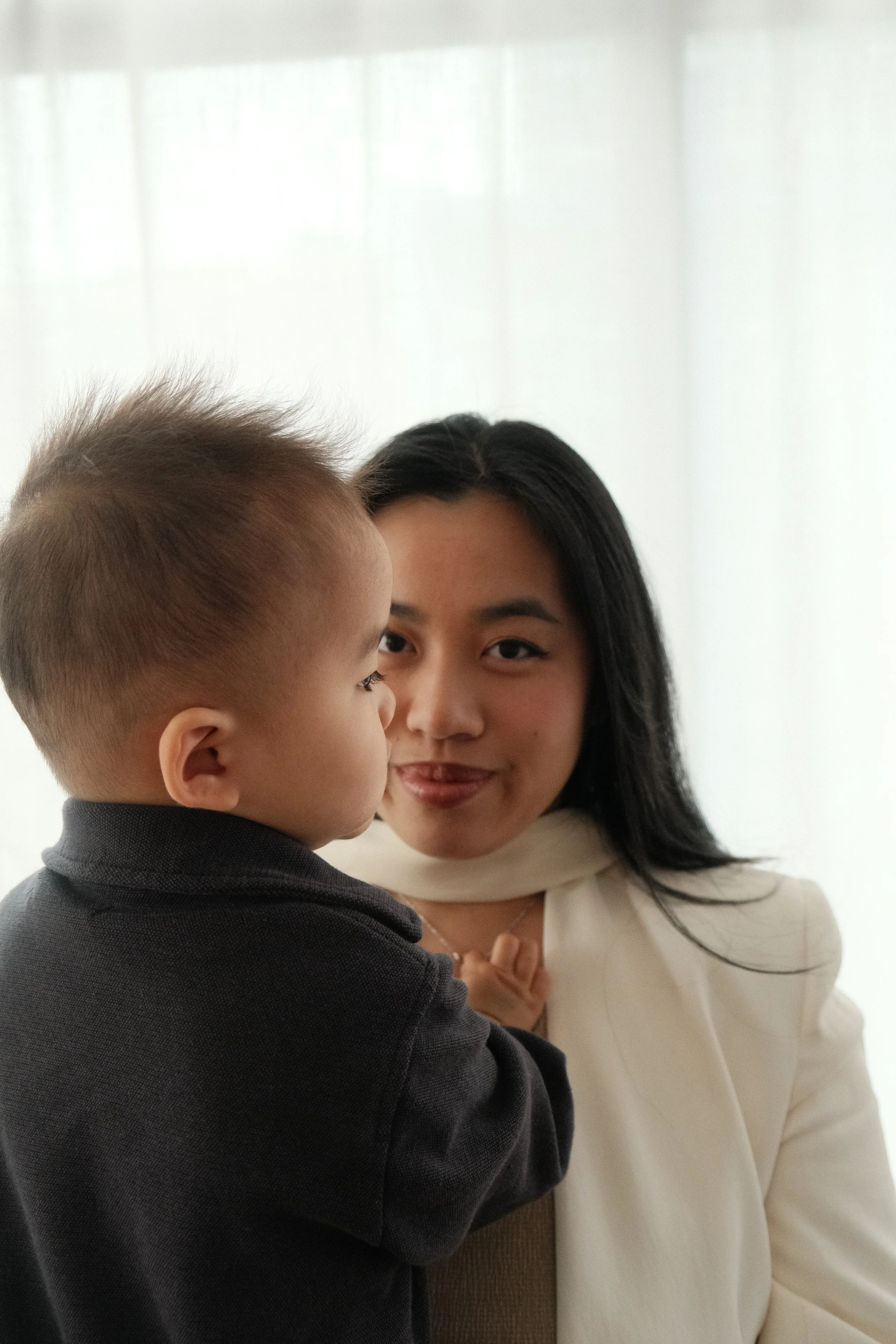 A woman with black hair and a beige blazer smiling while holding a young boy with short blonde hair in a gray sweater.