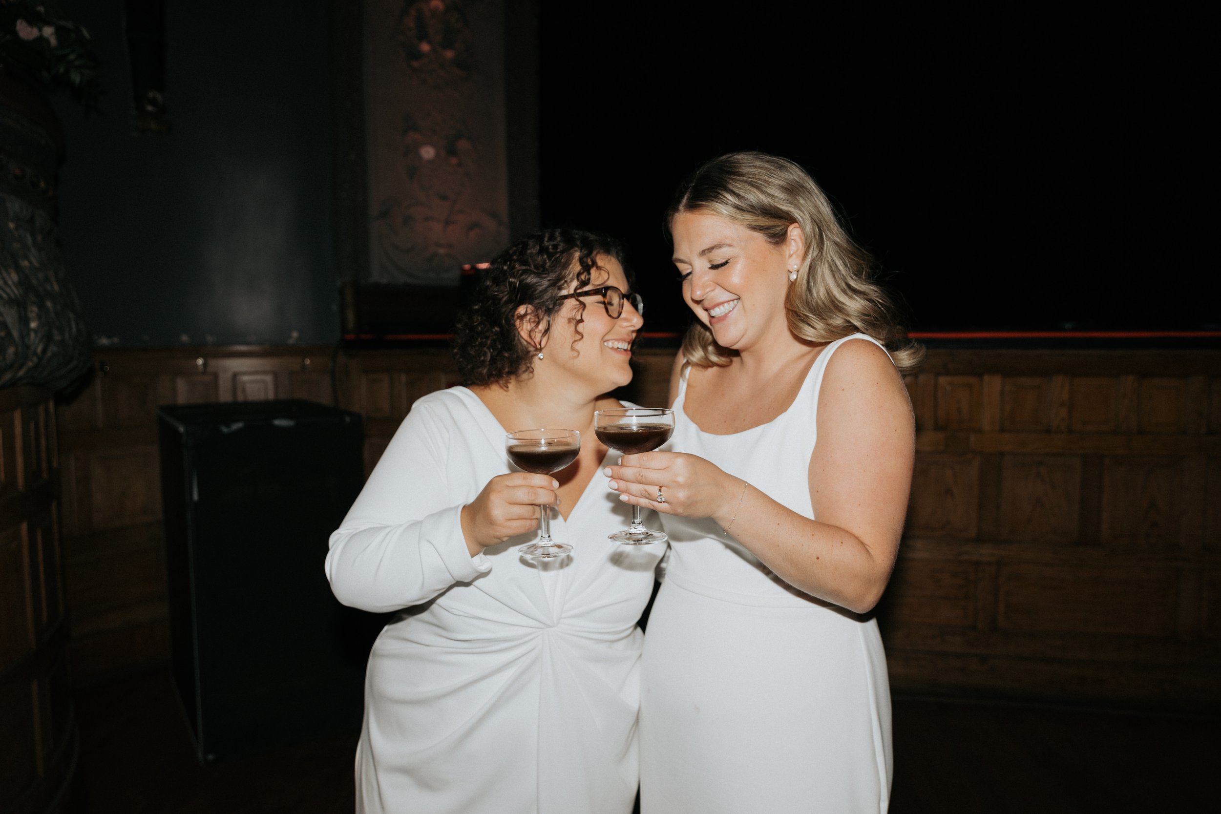 Two women in white dresses joyfully toasting with glasses of red wine indoors.