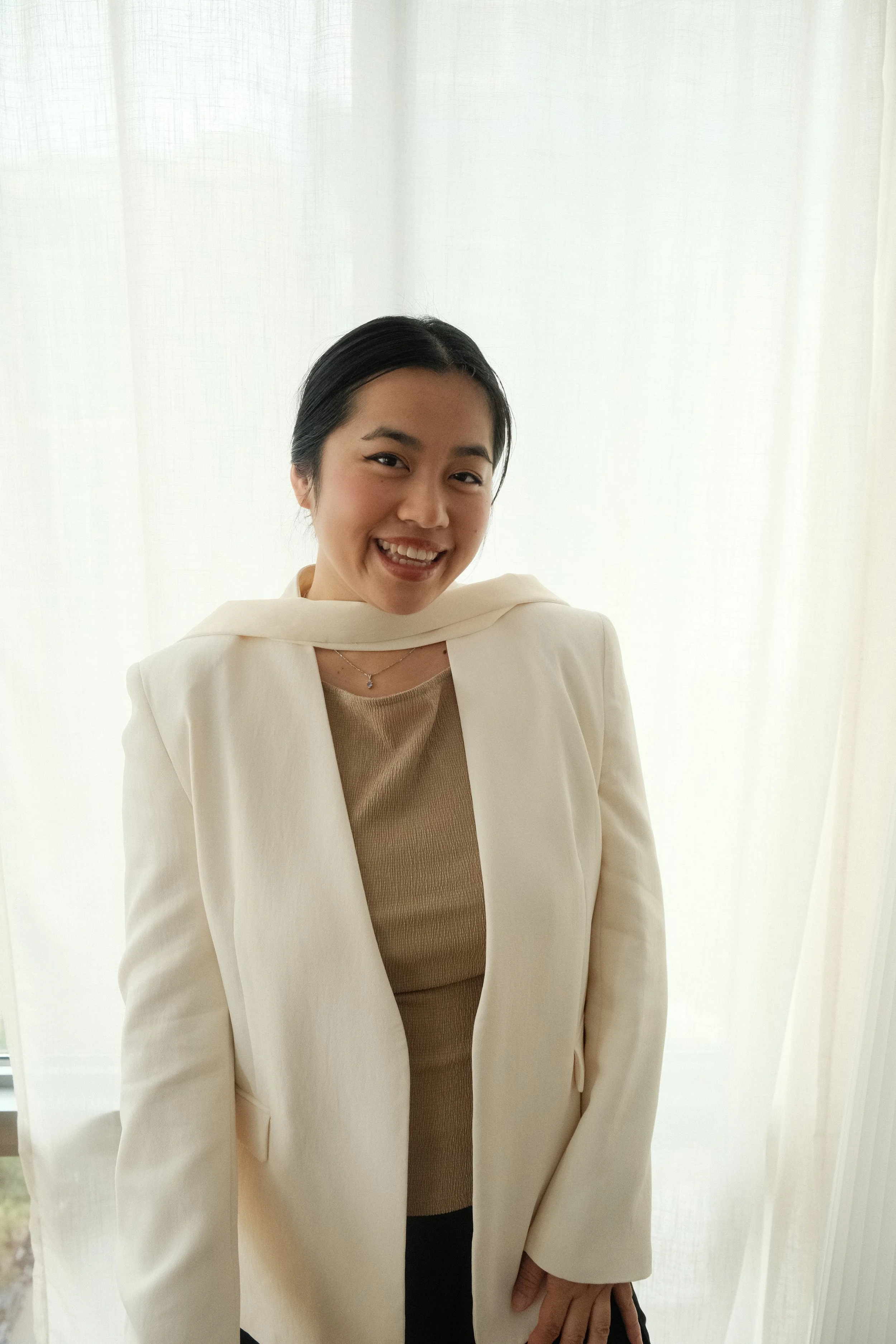A woman with dark hair smiling and wearing a beige top, cream blazer, and jewelry, standing in front of a white curtain.