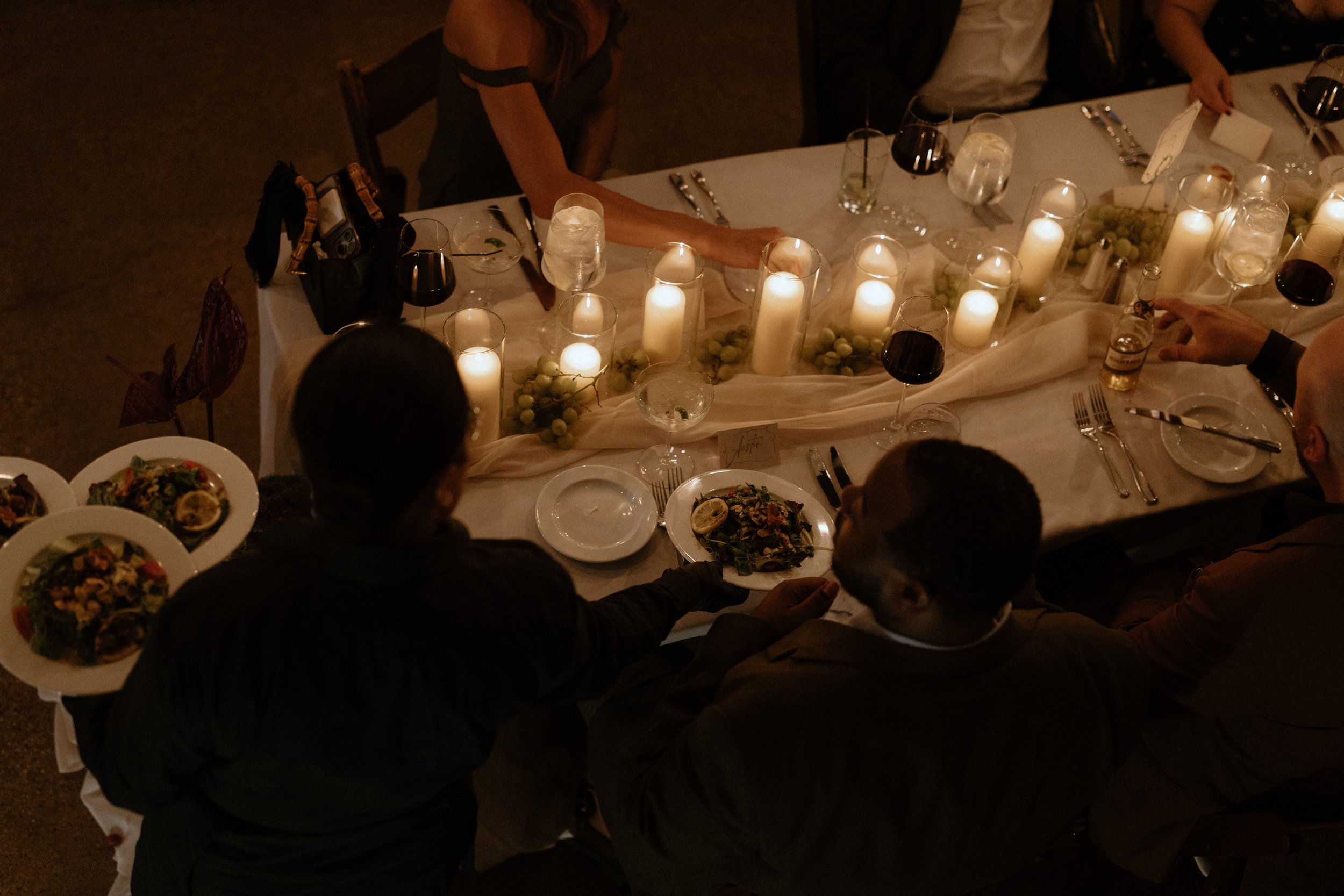 A dinner table decorated with white candles, green grapes, and elegant tableware, with guests serving themselves. The scene appears to be during the evening at a formal event.