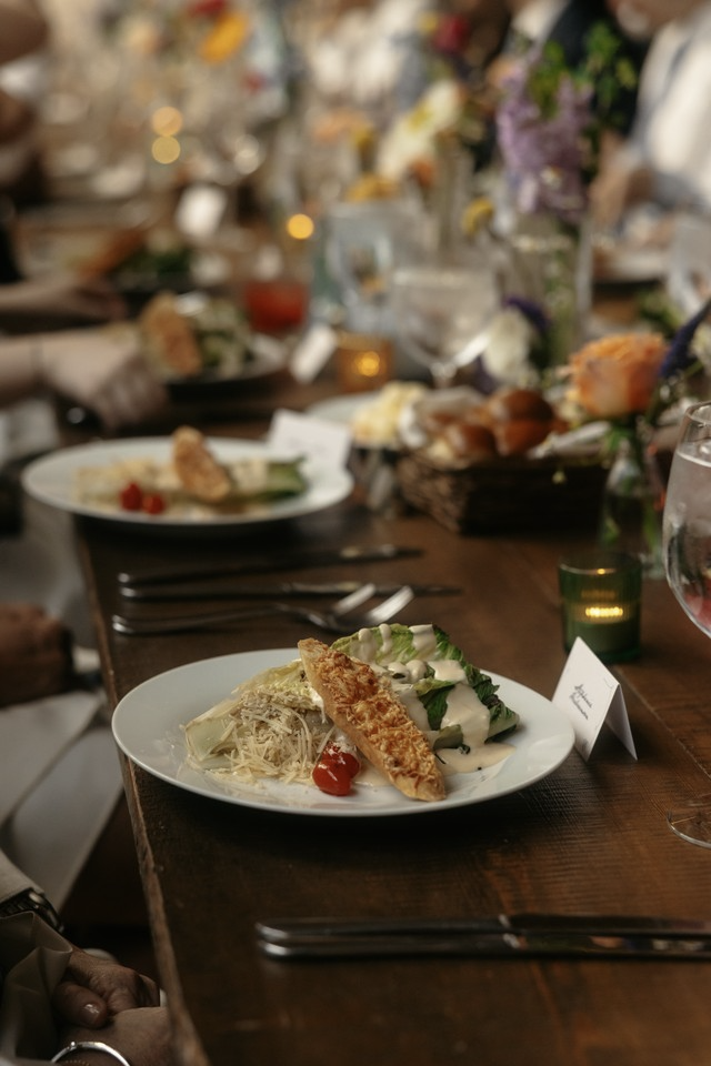 A dinner table with a plate of salad, breaded fried fish, and shredded cheese, surrounded by utensils, glasses, and floral centerpieces.