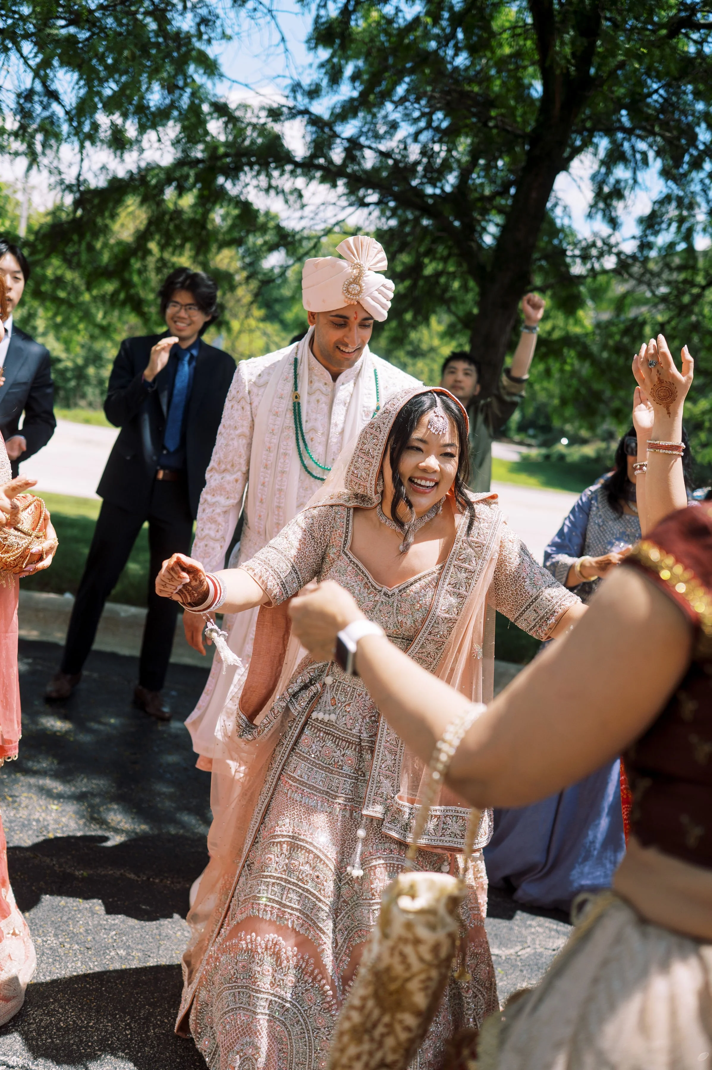 Indian bride and groom celebrating their wedding outdoors, with women in traditional attire, dancing and smiling under a large tree.
