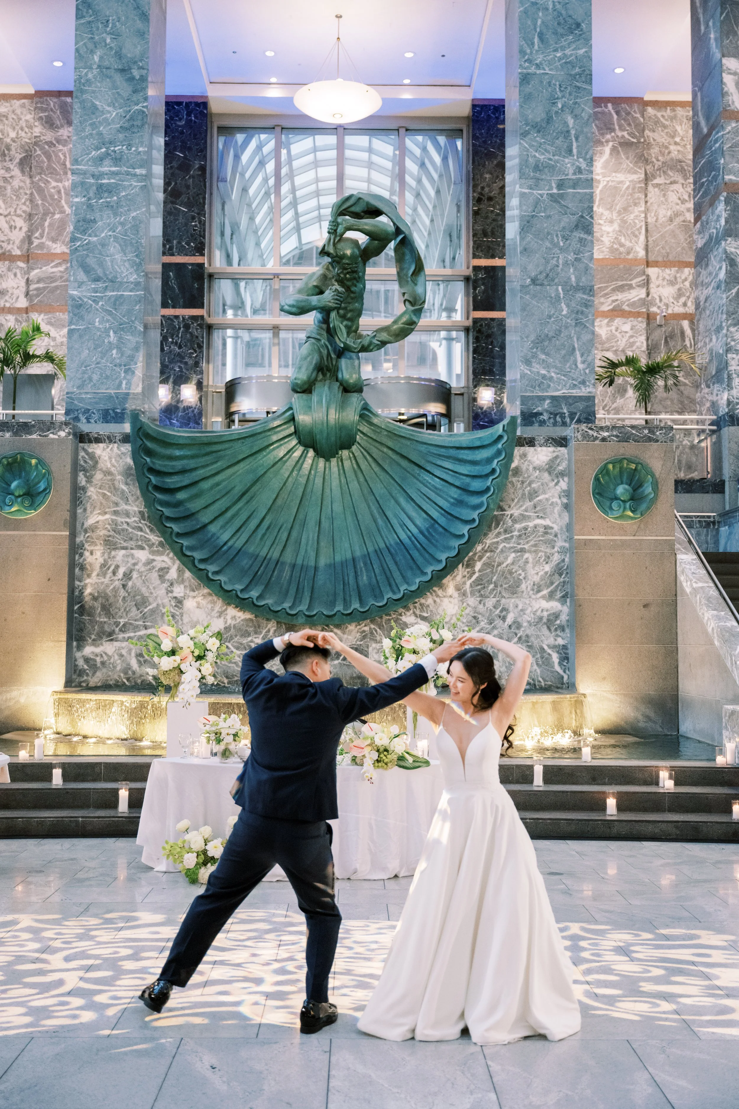 A couple dancing in a wedding reception inside a large, elegant building with marble walls, a statue of a mythological figure, and a decorated table with flowers in the background.