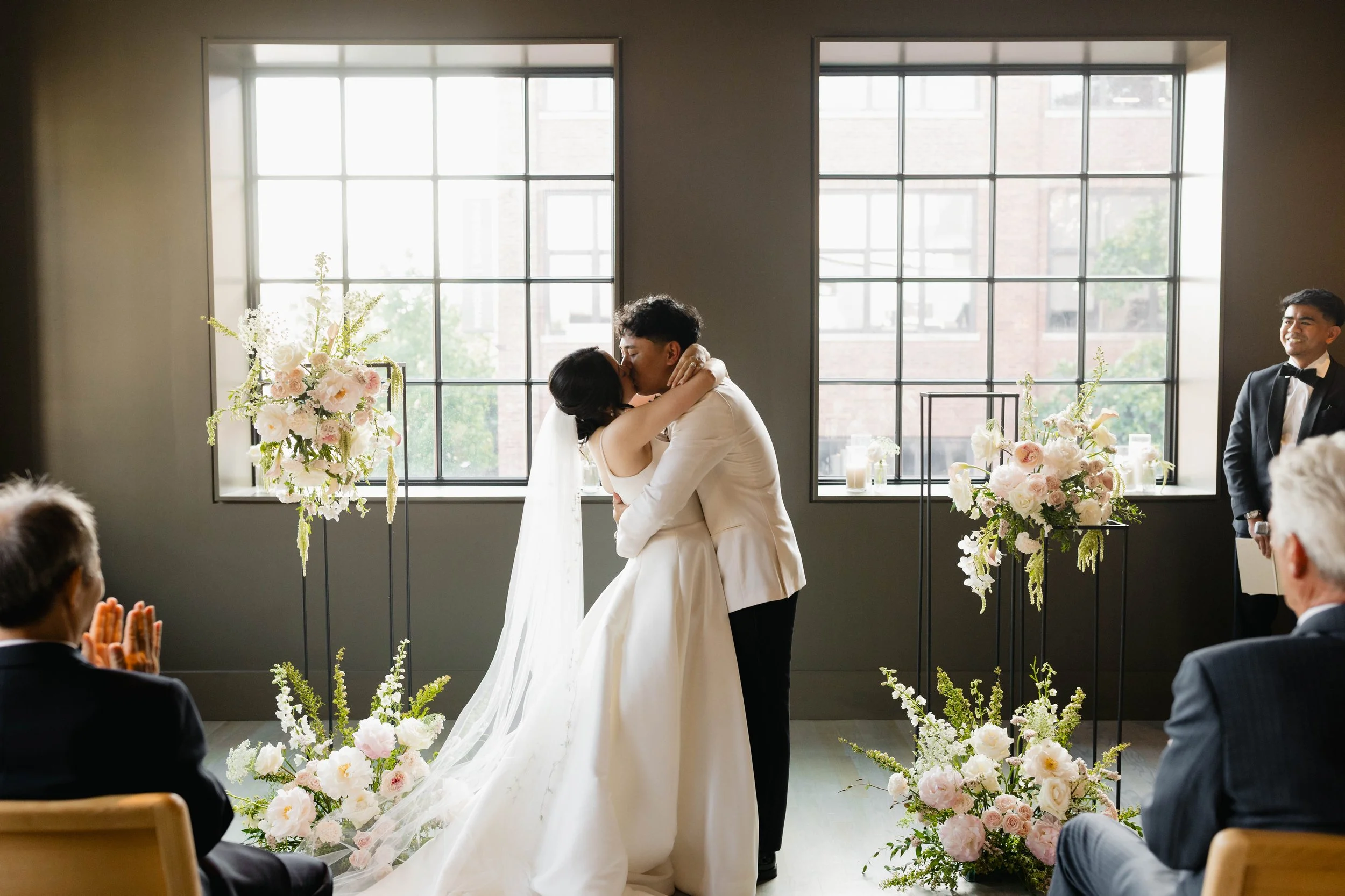 A bride and groom kiss during their wedding ceremony in front of tall windows with trees outside, decorated with floral arrangements and candles; a smiling officiant stands on the right, and the guests are clapping.