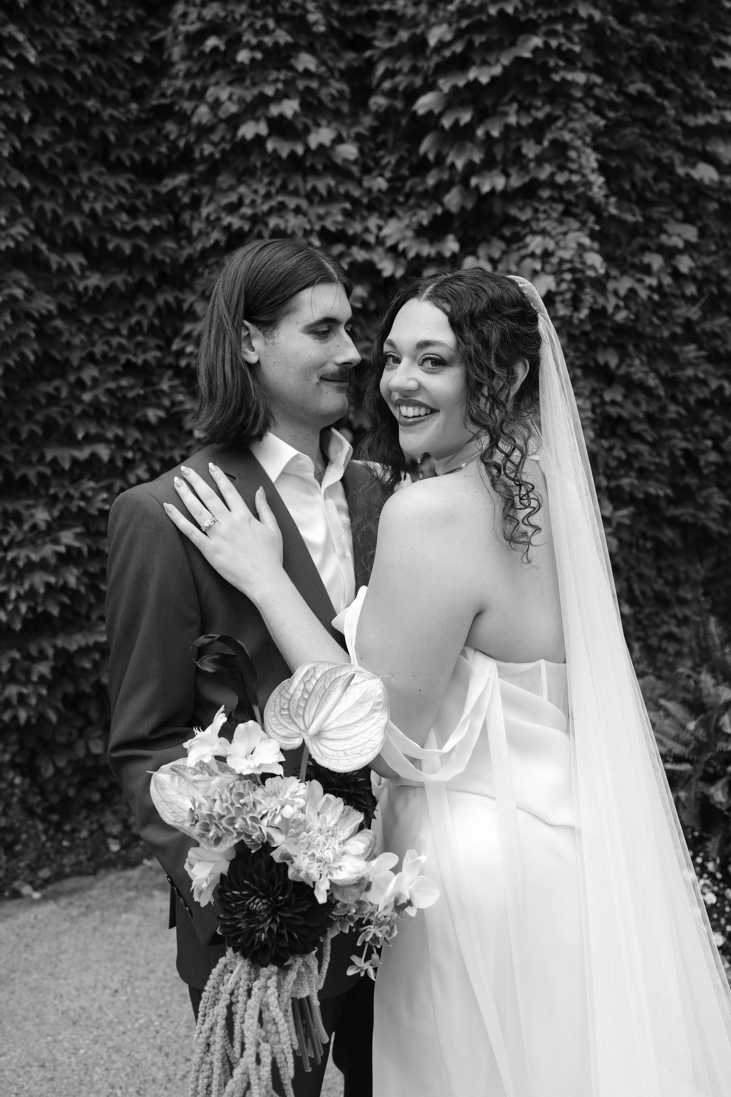 A black-and-white photo of a wedding couple, with the woman in a strapless wedding dress and veil, and the man in a suit, standing close together outdoors in front of a wall of dense foliage, smiling.