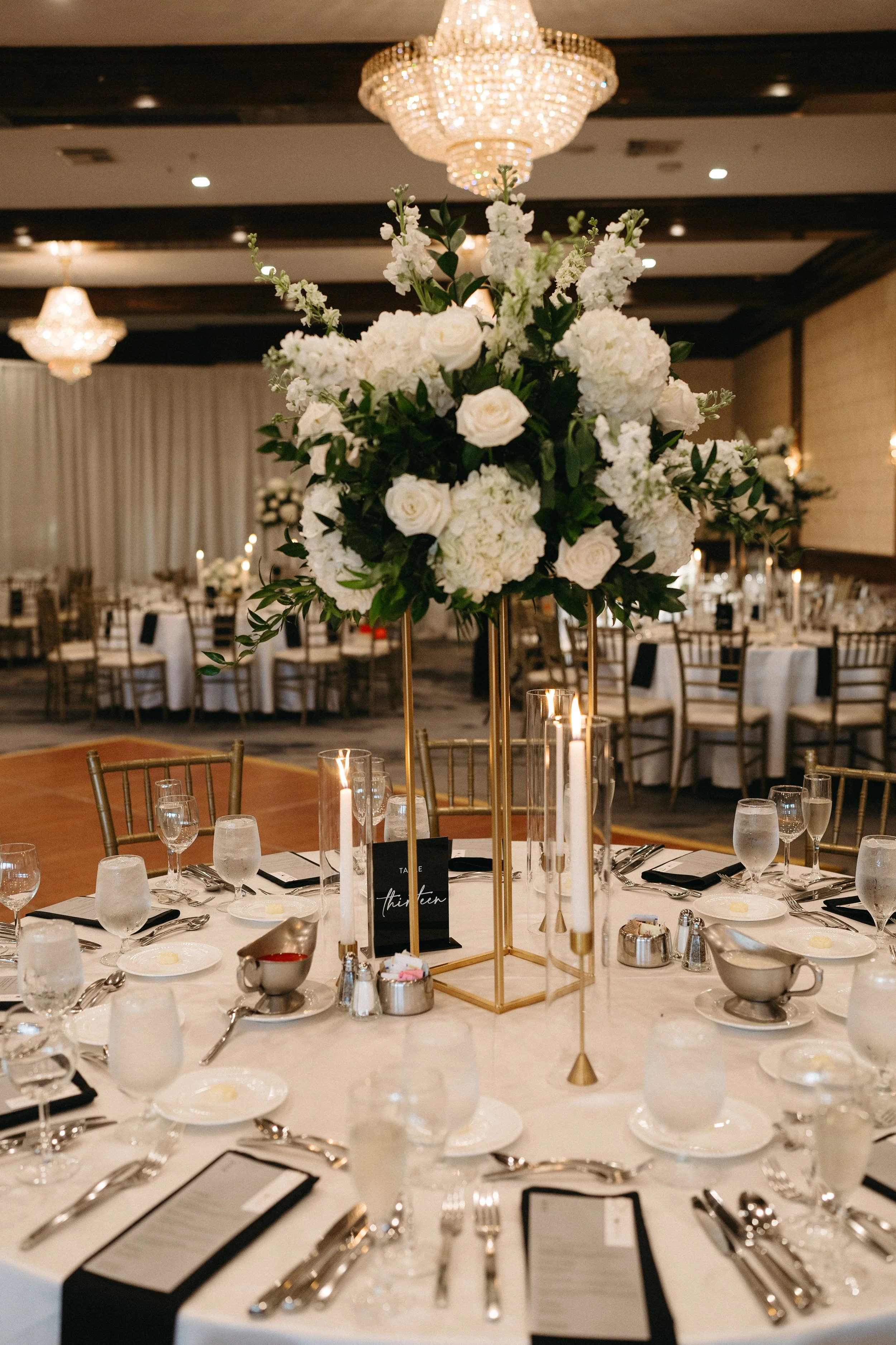 A formal banquet table setup with a tall floral centerpiece of white flowers and green leaves, surrounded by candles, glassware, plates, silverware, and black menus with white writing in a decorated banquet hall with chandeliers.