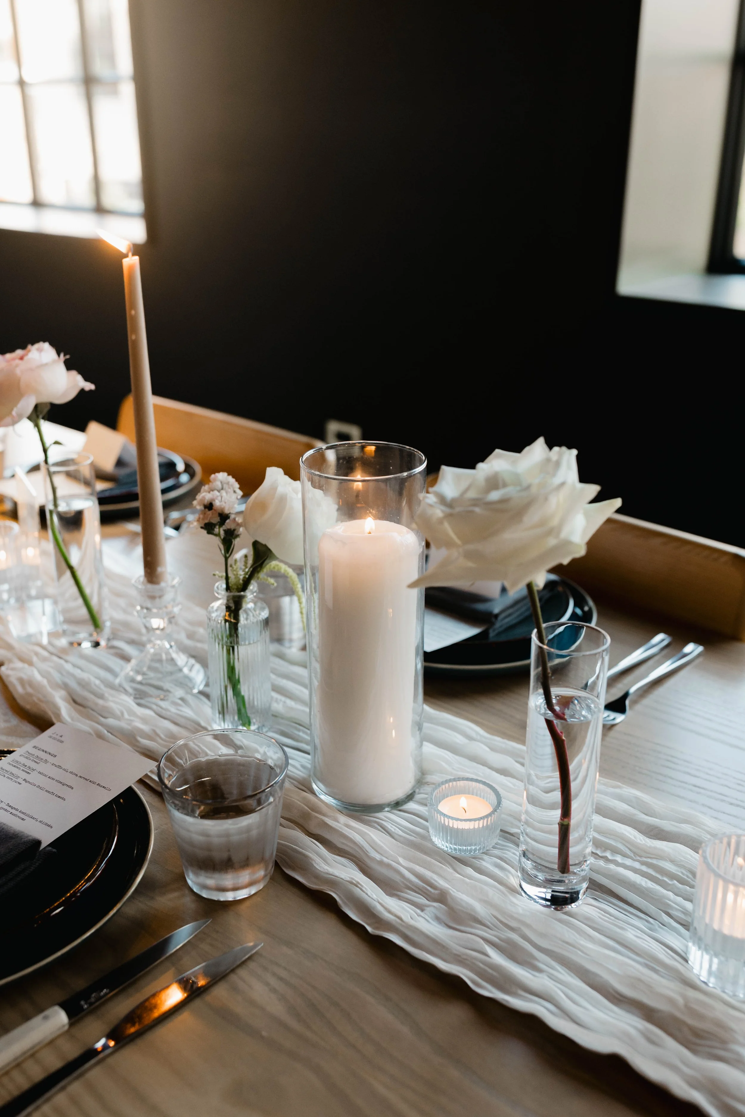 A table set for a dinner with candles, flowers, and black plates and silverware.