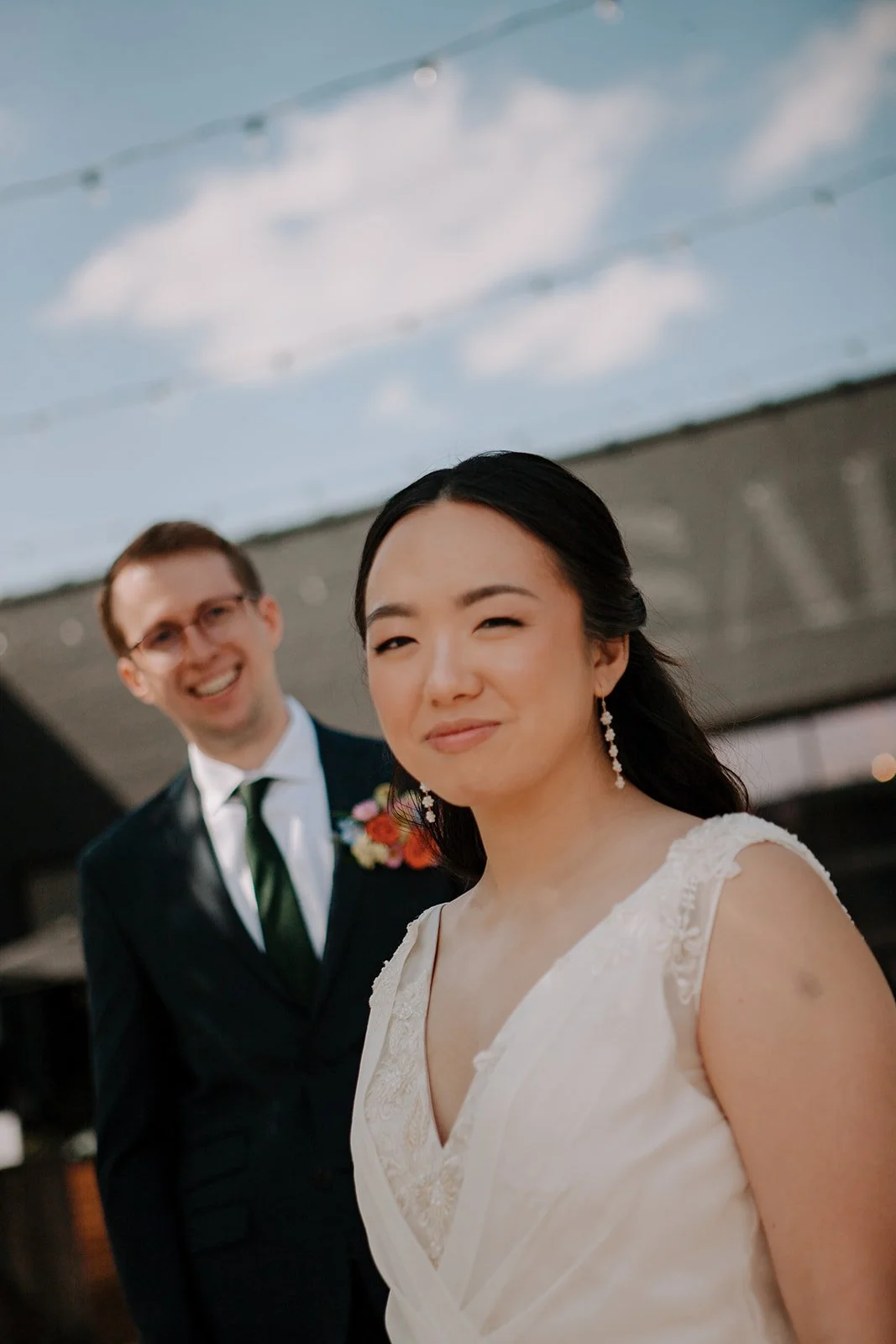 A woman in a white dress winks at the camera while a man in a suit with glasses and a boutonniere stands smiling in the background during an outdoor event under a partly cloudy sky.