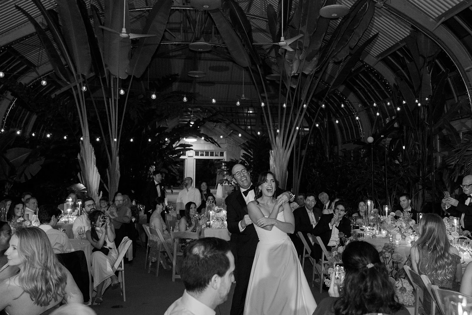 A black and white photo of a wedding reception with a bride and groom dancing among seated guests in an elegant venue with tropical plants and string lights overhead.