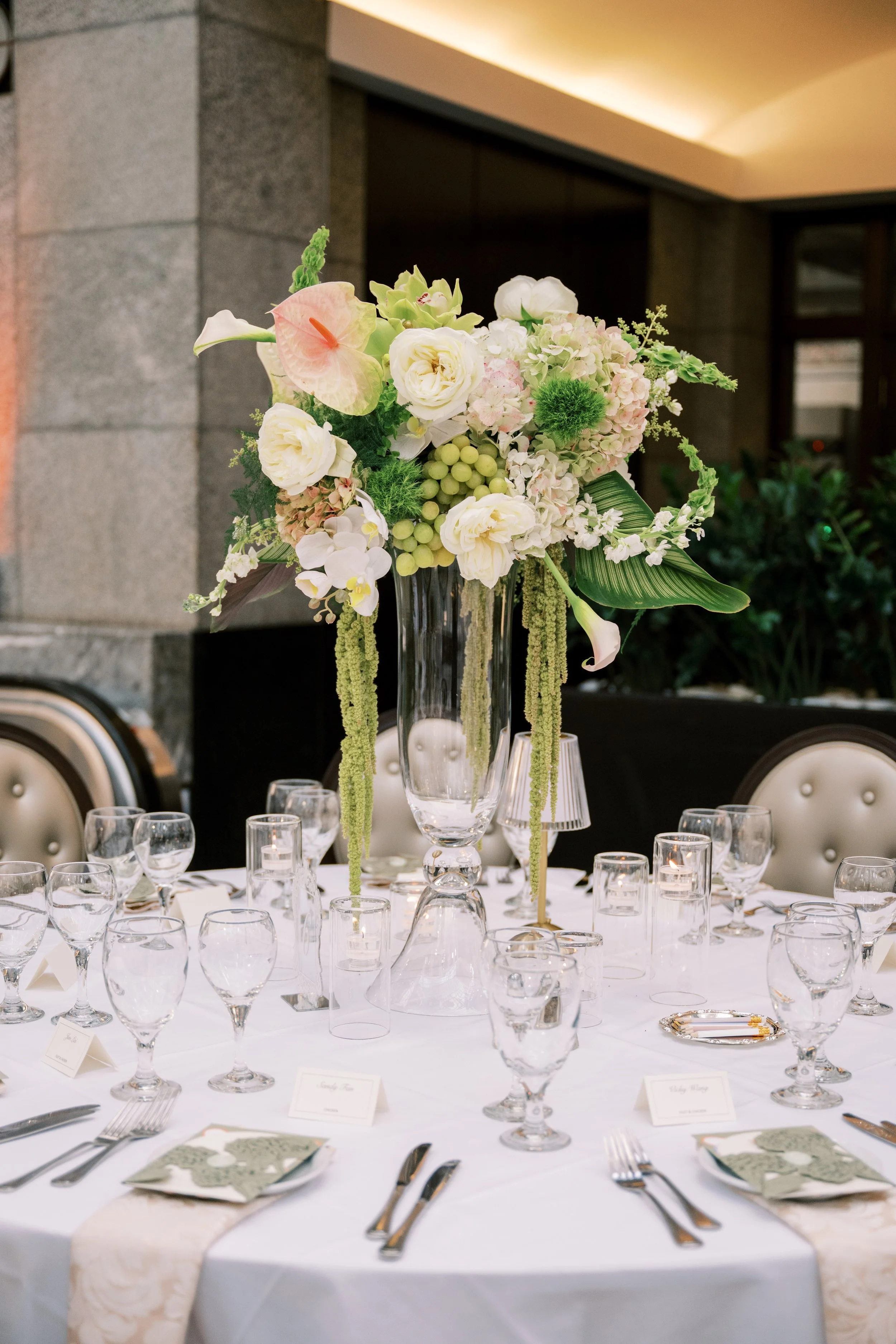 Elegant dining table with a large floral centerpiece in a tall glass vase, surrounded by multiple wine glasses and utensils on a white tablecloth, set in a formal event setting.