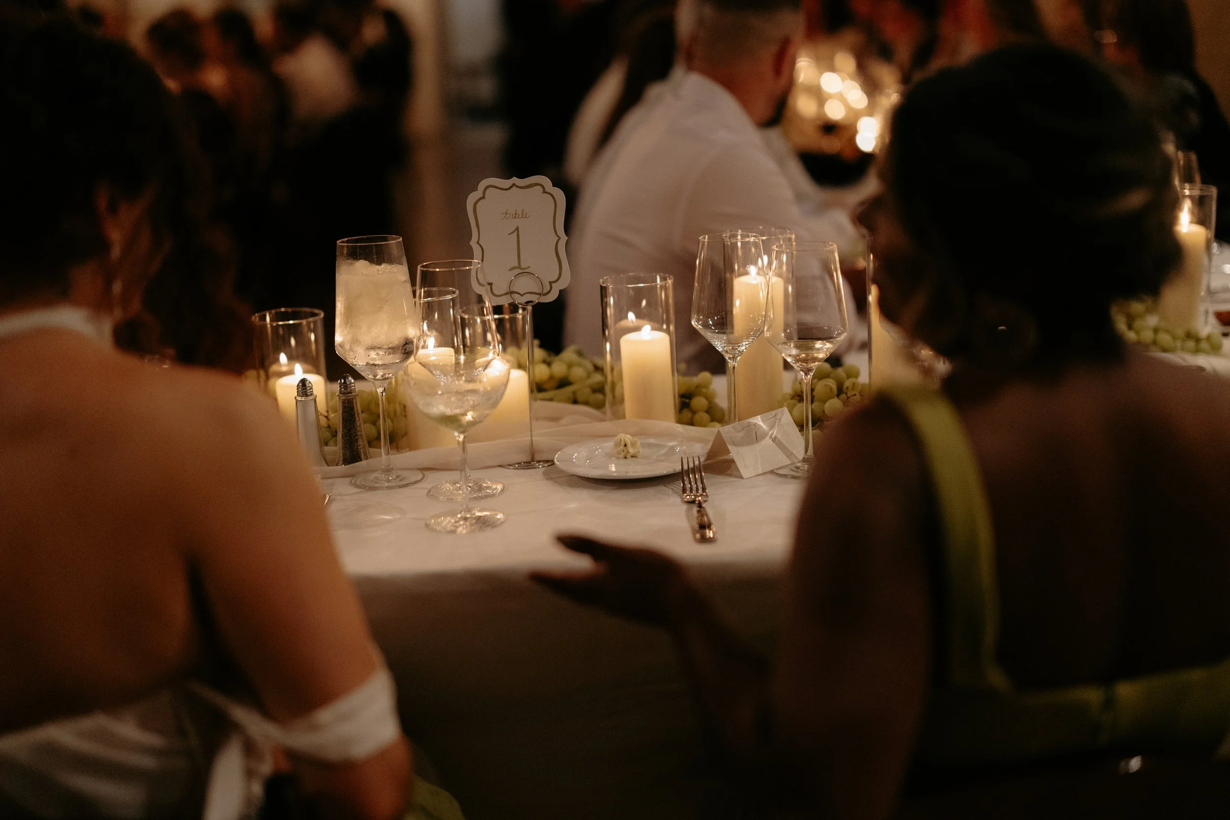 A wedding reception table with candles and a sign indicating table 1, blurred figures of wedding guests in formal attire in the background.