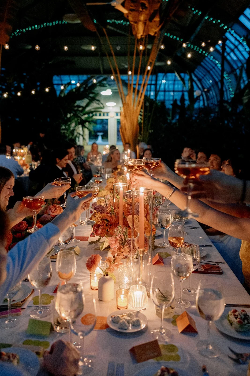 People celebrating at a dinner party, raising glasses in a toast around a long table with candles, floral centerpieces, and tableware inside a decorated venue with string lights and a glass ceiling.