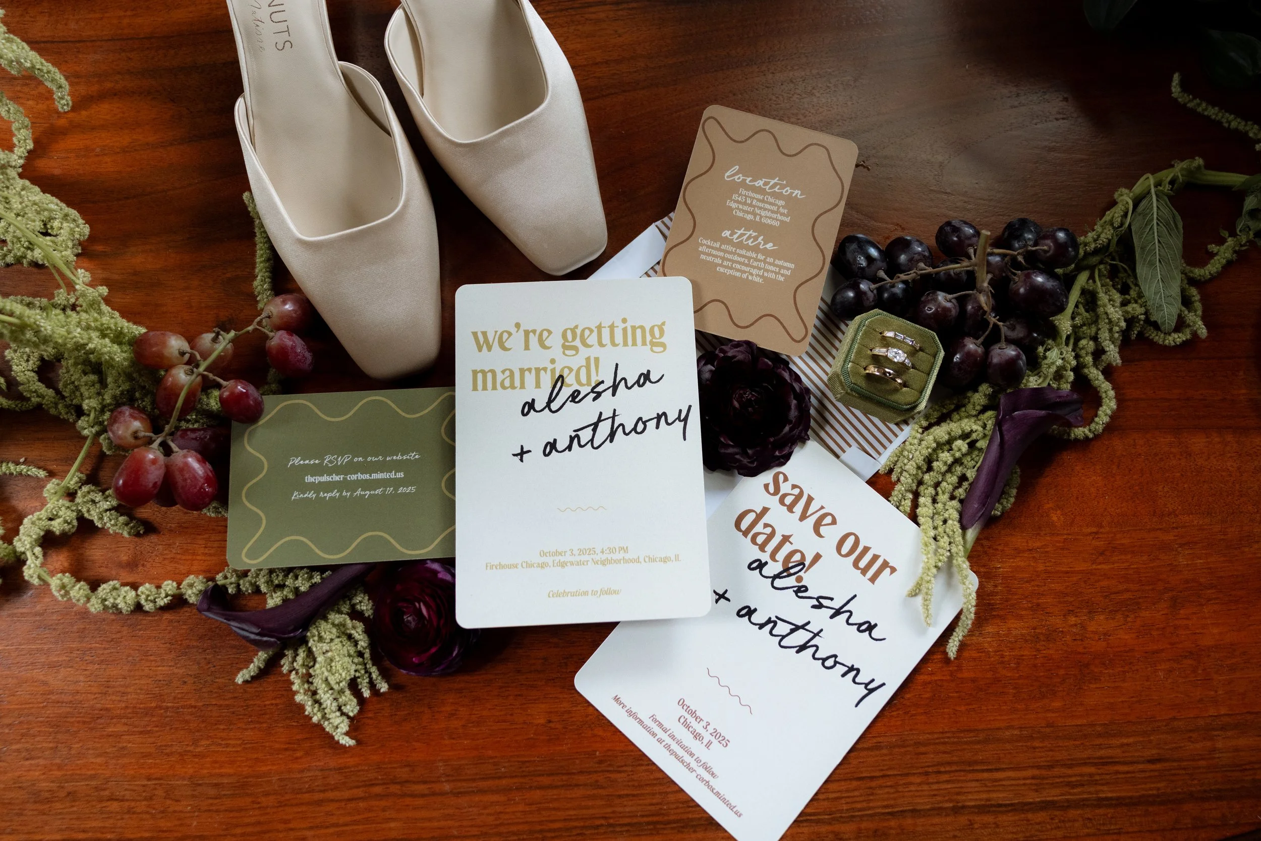 A pair of beige satin wedding shoes surrounded by wedding invitations, grapes, and greenery on a wooden surface.