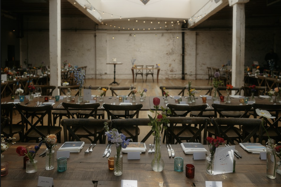 Long wedding reception table decorated with flowers, candles, and place settings in an industrial-style venue with string lights and a small dance area in the background.