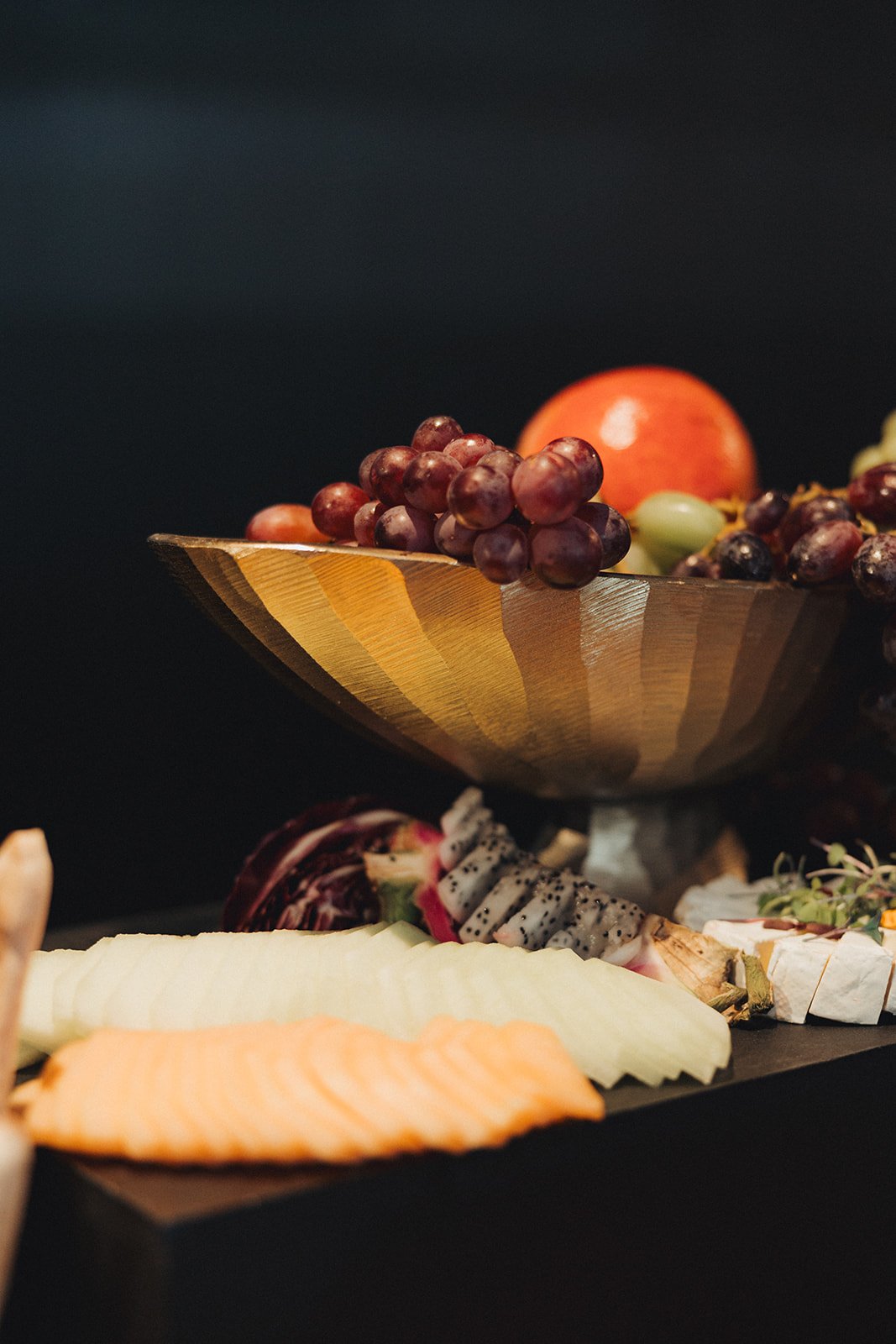 Close-up of a wooden bowl filled with grapes and an orange, with a variety of sliced fruits including melons, dragon fruit, and possibly cheese on a black surface.