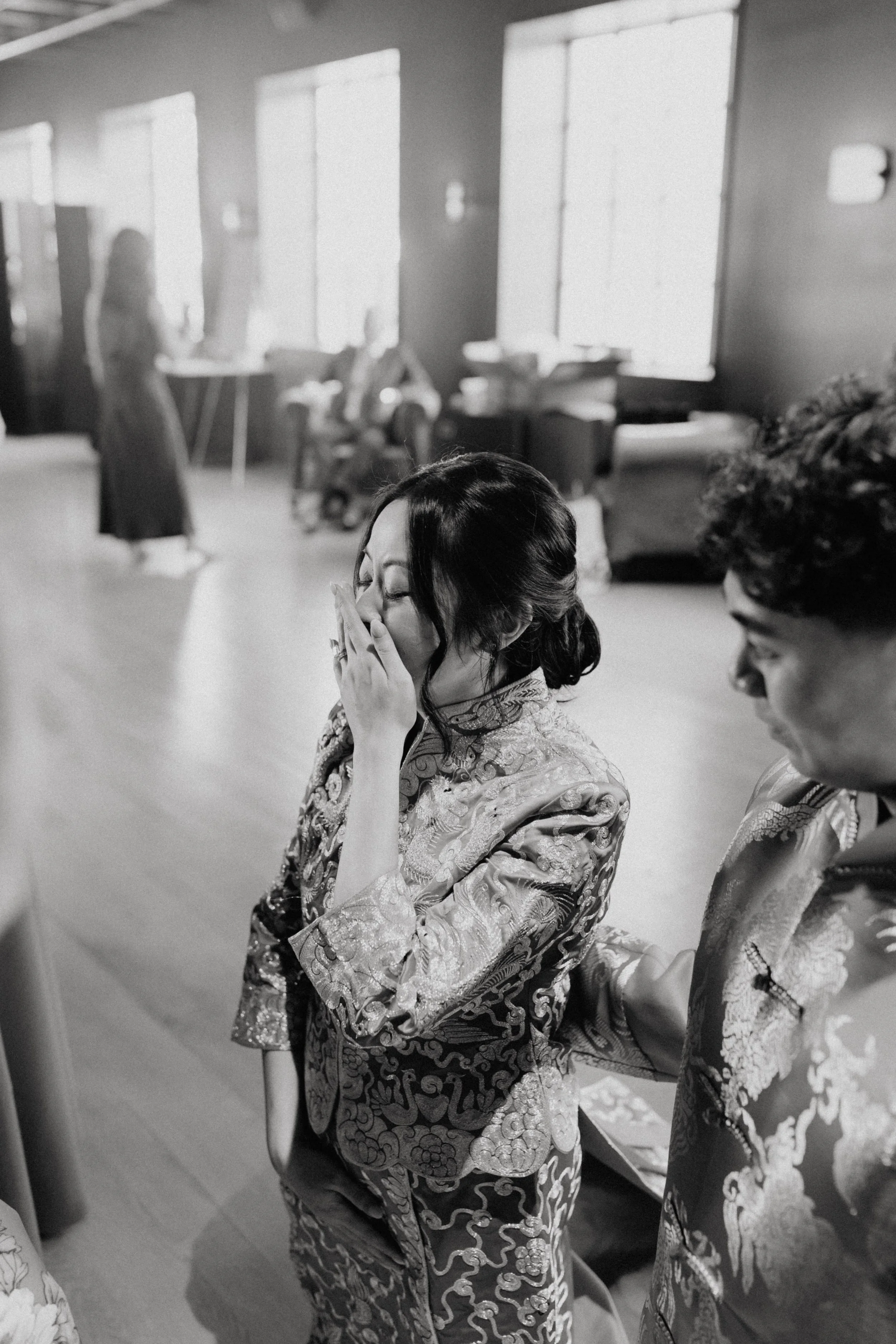 Two women in traditional ornate outfits are seated, one bows her head and closes her eyes with her hand near her face, in what appears to be a ceremonial or cultural setting, with other people and large windows in the background.