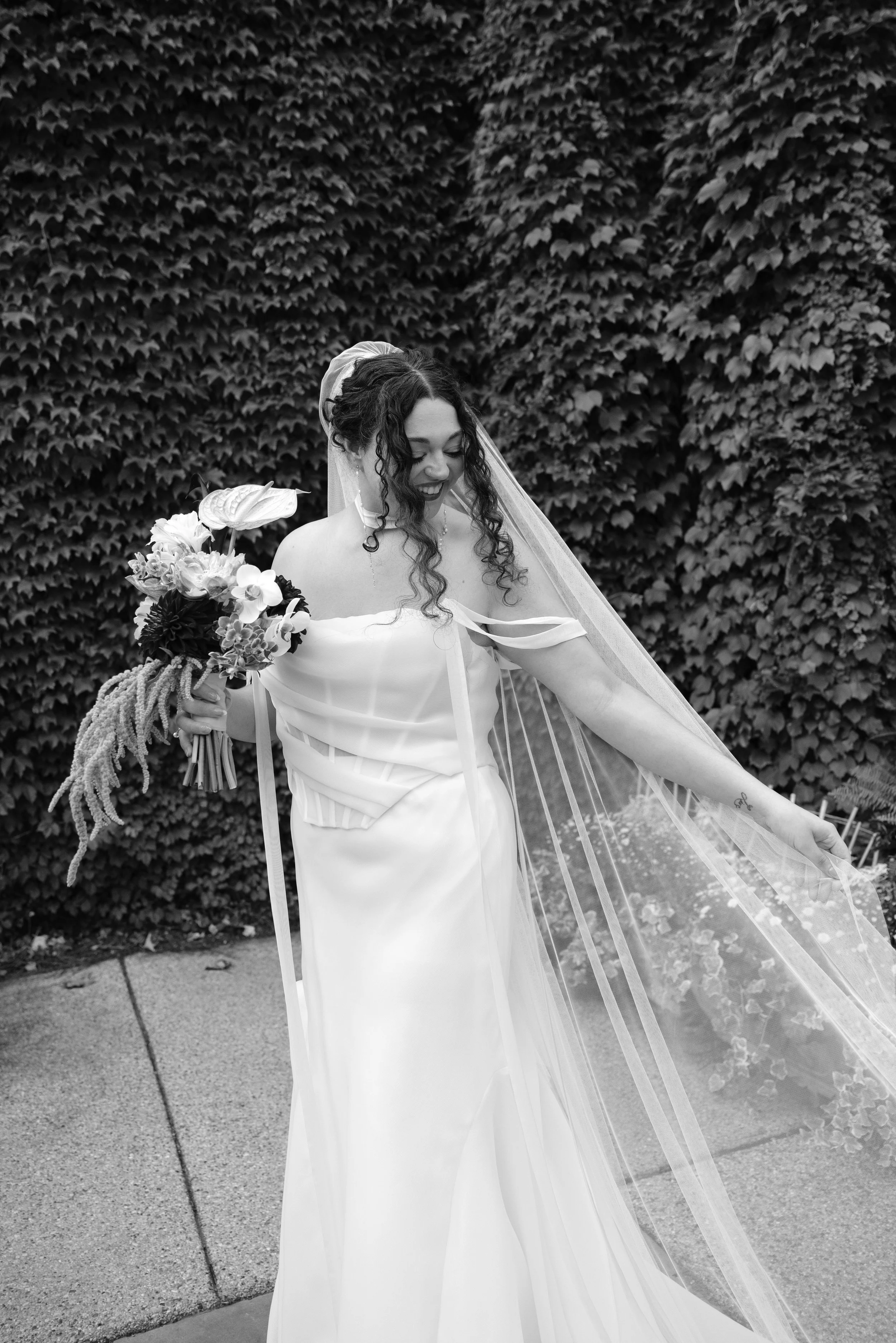 A woman in a graduation gown and veil holding a bouquet of flowers, standing outdoors in front of a wall covered in vines.