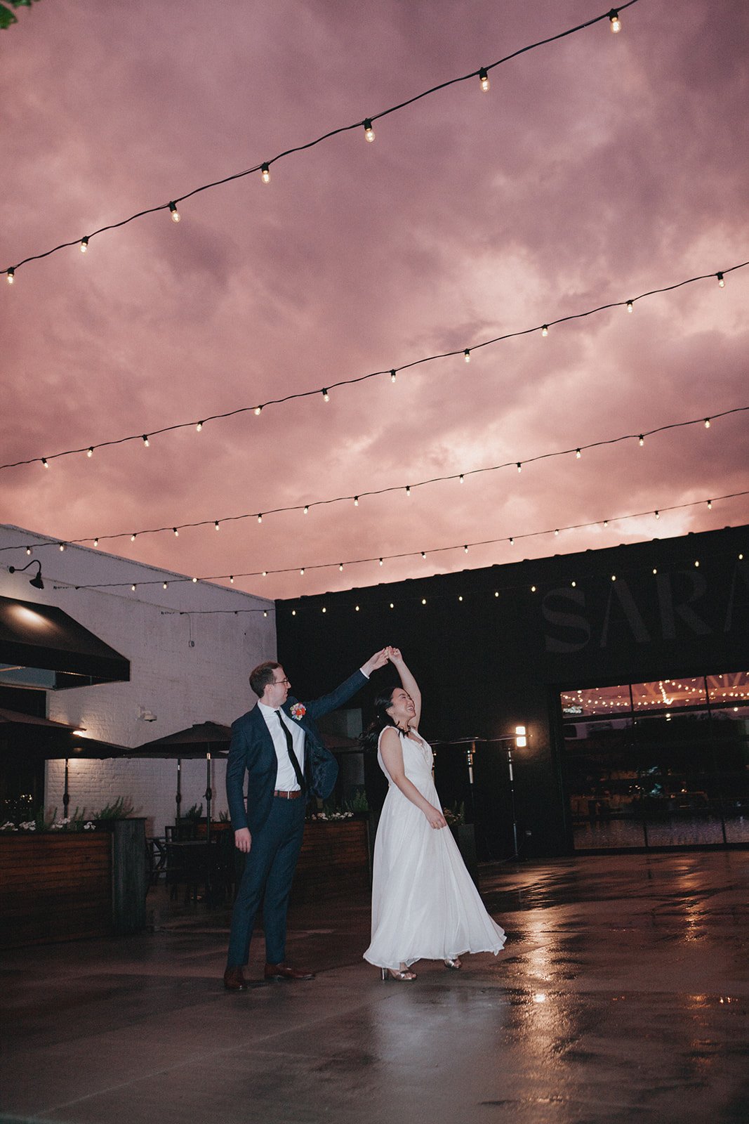 A couple dancing outdoors at night under string lights with a pink cloudy sky.
