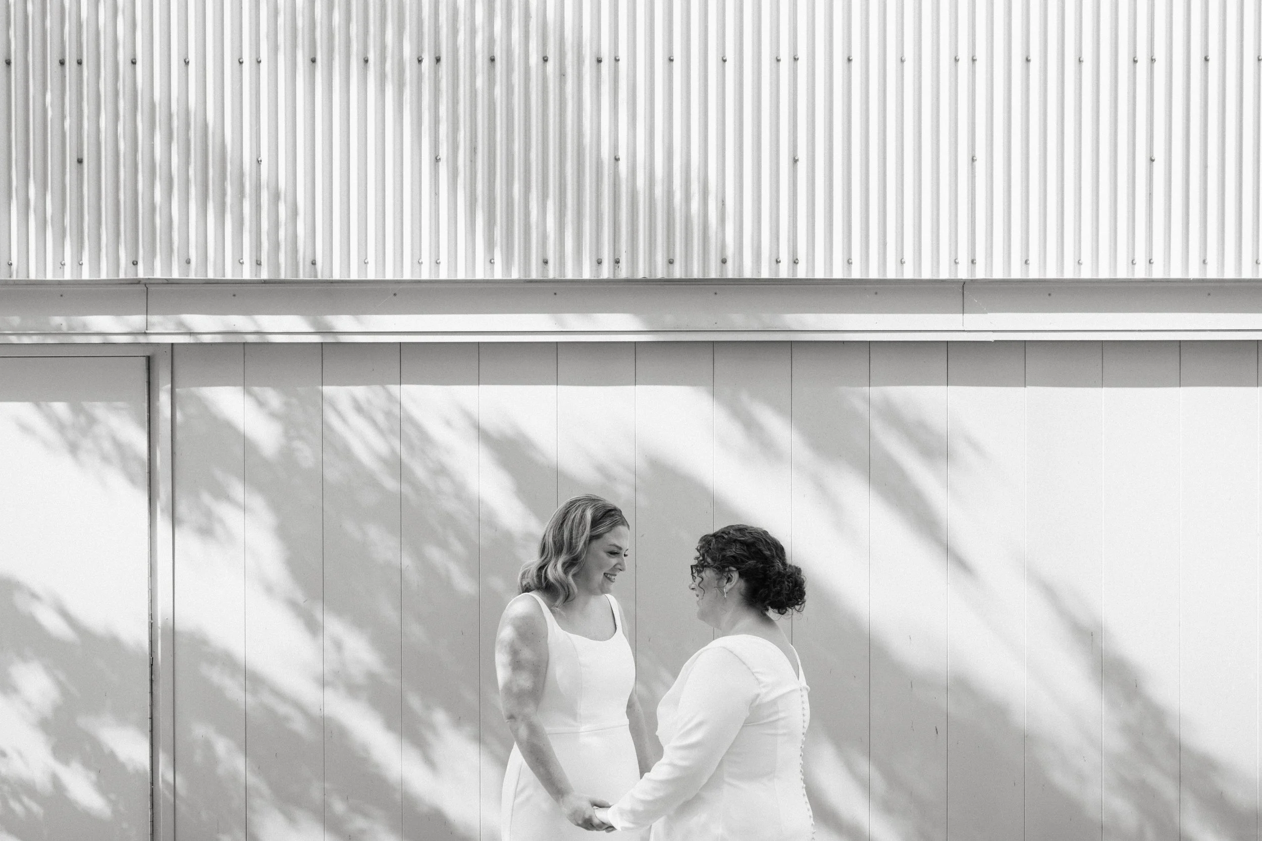 Two women, one with blonde hair and the other with curly hair, hold hands and smile at each other outdoors against a white wall with shadows of tree branches.