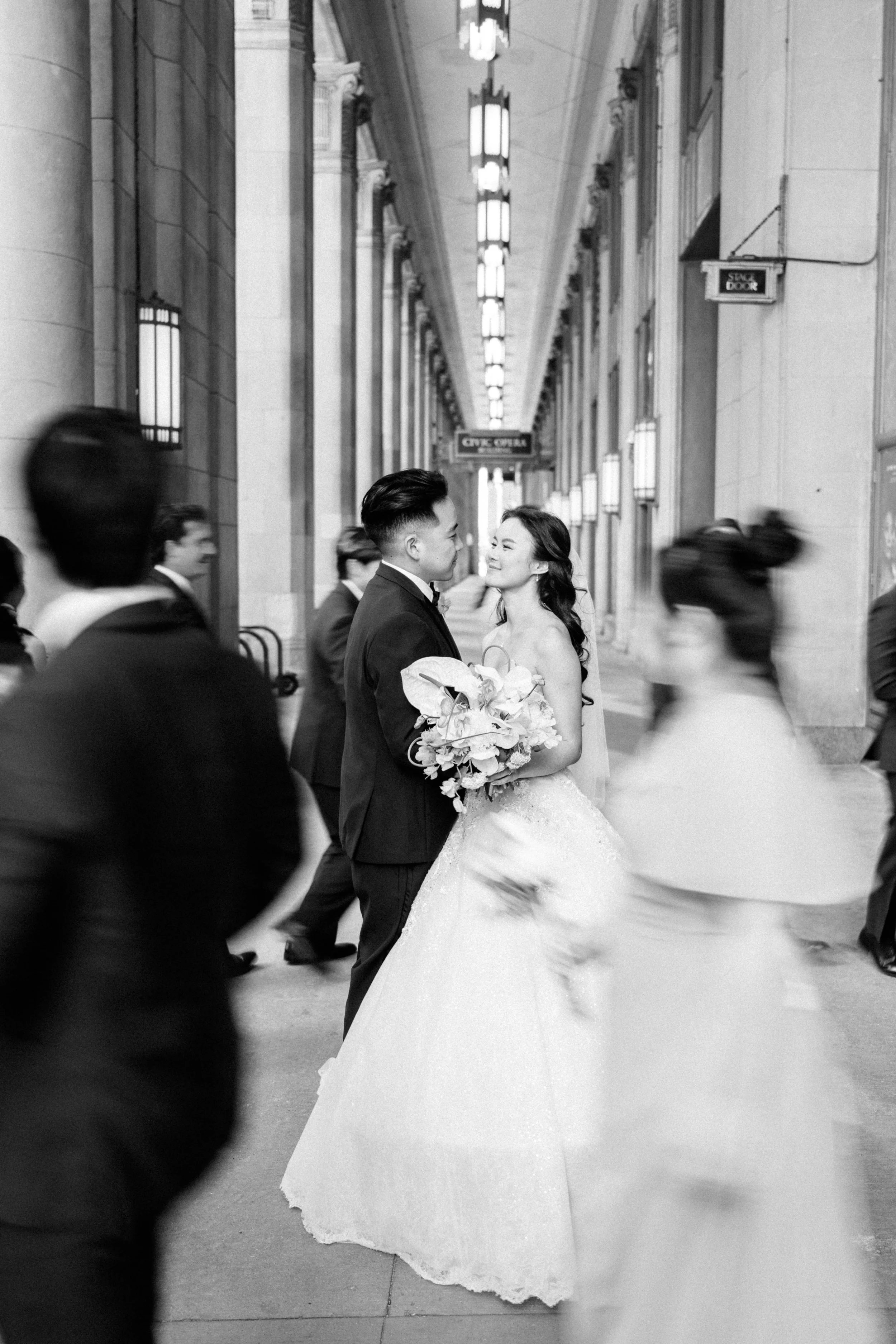 A bride and groom dancing in a hallway with architectural details, lights, and a sign that says 'Serve Door.' The bride is holding a bouquet and smiling at the groom, who is dressed in a tuxedo.