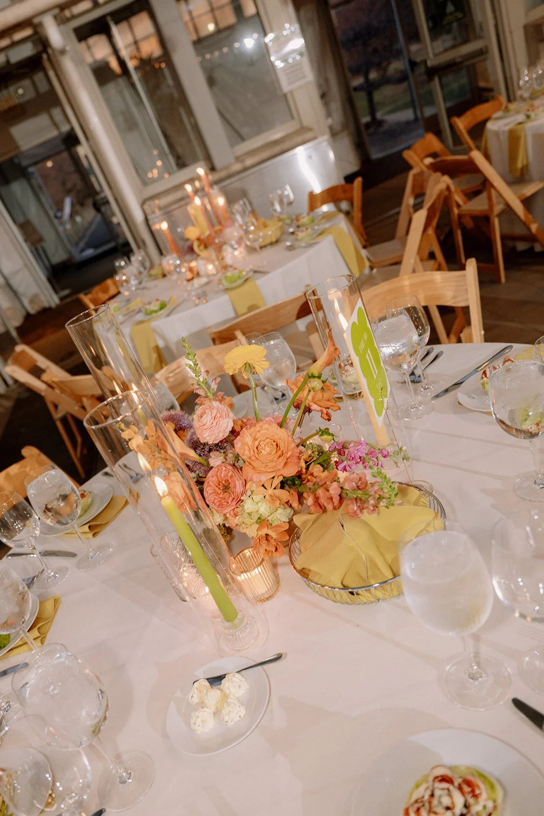 A banquet table decorated with pink and yellow flowers, candles in glass holders, tableware, and a small card.