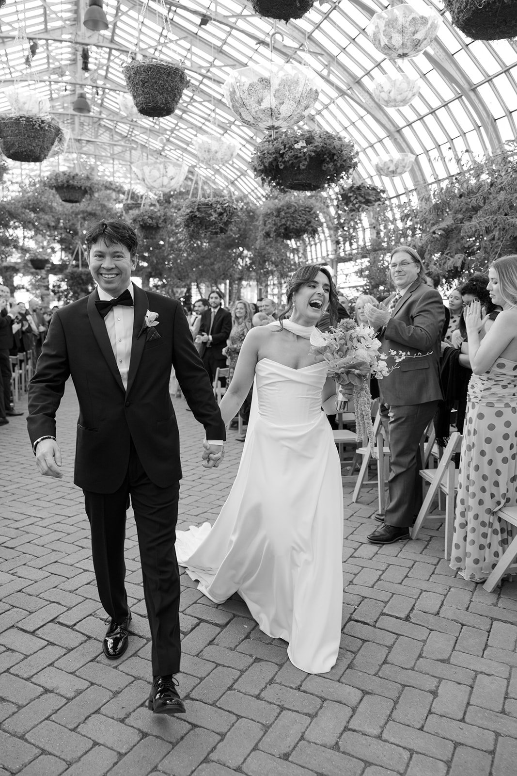 A newlywed couple holding hands, smiling, and walking down the aisle of a greenhouse wedding venue with guests clapping and celebrating around them.